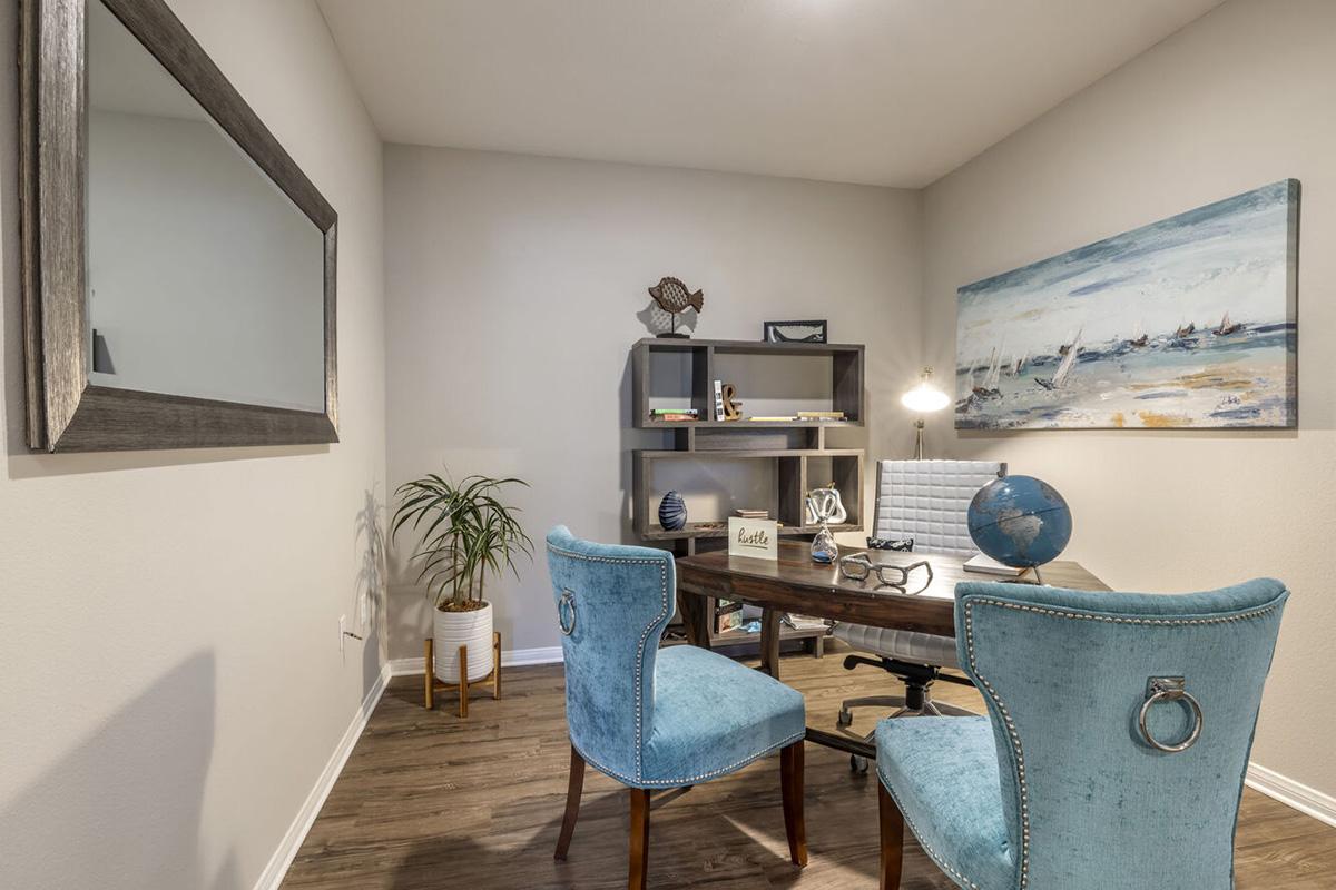 A cozy home office featuring a wooden desk with a globe, a desk lamp, and decorative items on a shelf. Two aqua-blue upholstered chairs are positioned at the desk. A plant in a white pot adds a touch of greenery, while a large mirror enhances the room's space. The walls are painted in neutral tones.