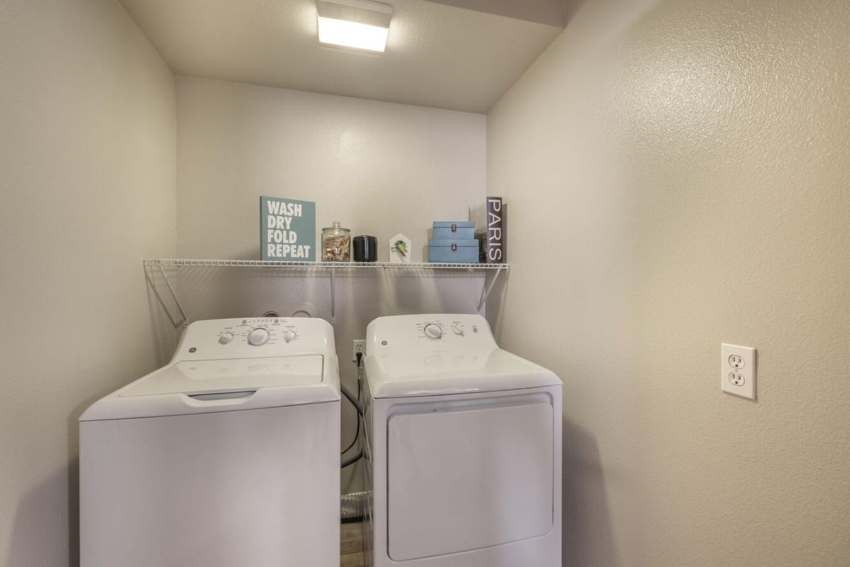 A clean and modern laundry room featuring a pair of white washing machines side by side, a light gray wall, and a shelf above. The shelf holds decorative items, including a sign that reads "WASH DRY FOLD REPEAT," a small blue box, candles, and a tall decorative piece labeled "PARIS."