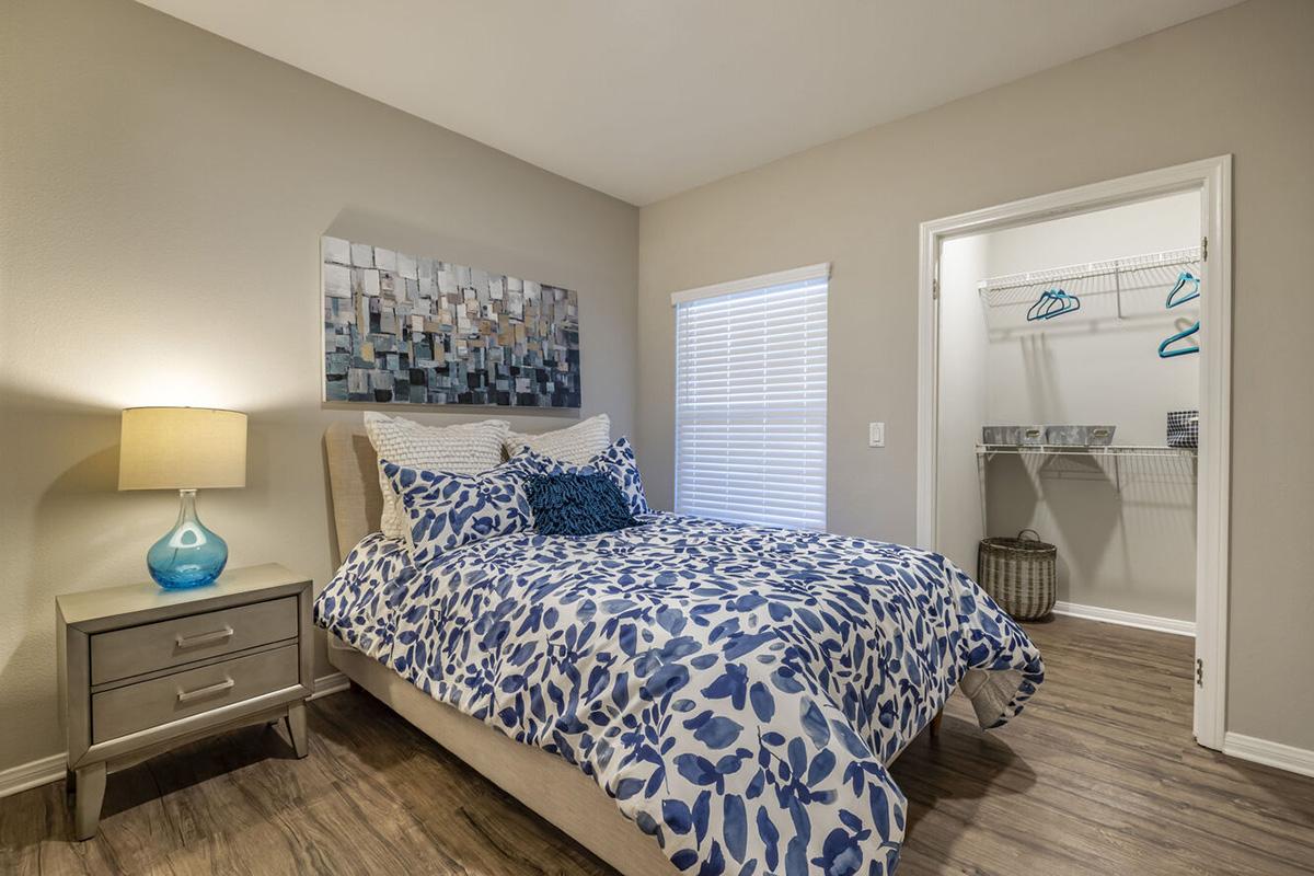 A cozy bedroom featuring a bed with a blue and white floral duvet cover, flanked by a nightstand with a decorative lamp. A piece of abstract wall art hangs above the bed. Natural light enters through a window, and a closet with hangers is visible in the background, along with a woven basket.