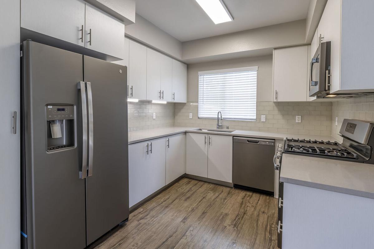 Modern kitchen with stainless steel appliances including a refrigerator, dishwasher, and stove. The cabinets are light-colored, and there's a window with blinds allowing natural light. The flooring is wood-like laminate, creating a contemporary and clean aesthetic.