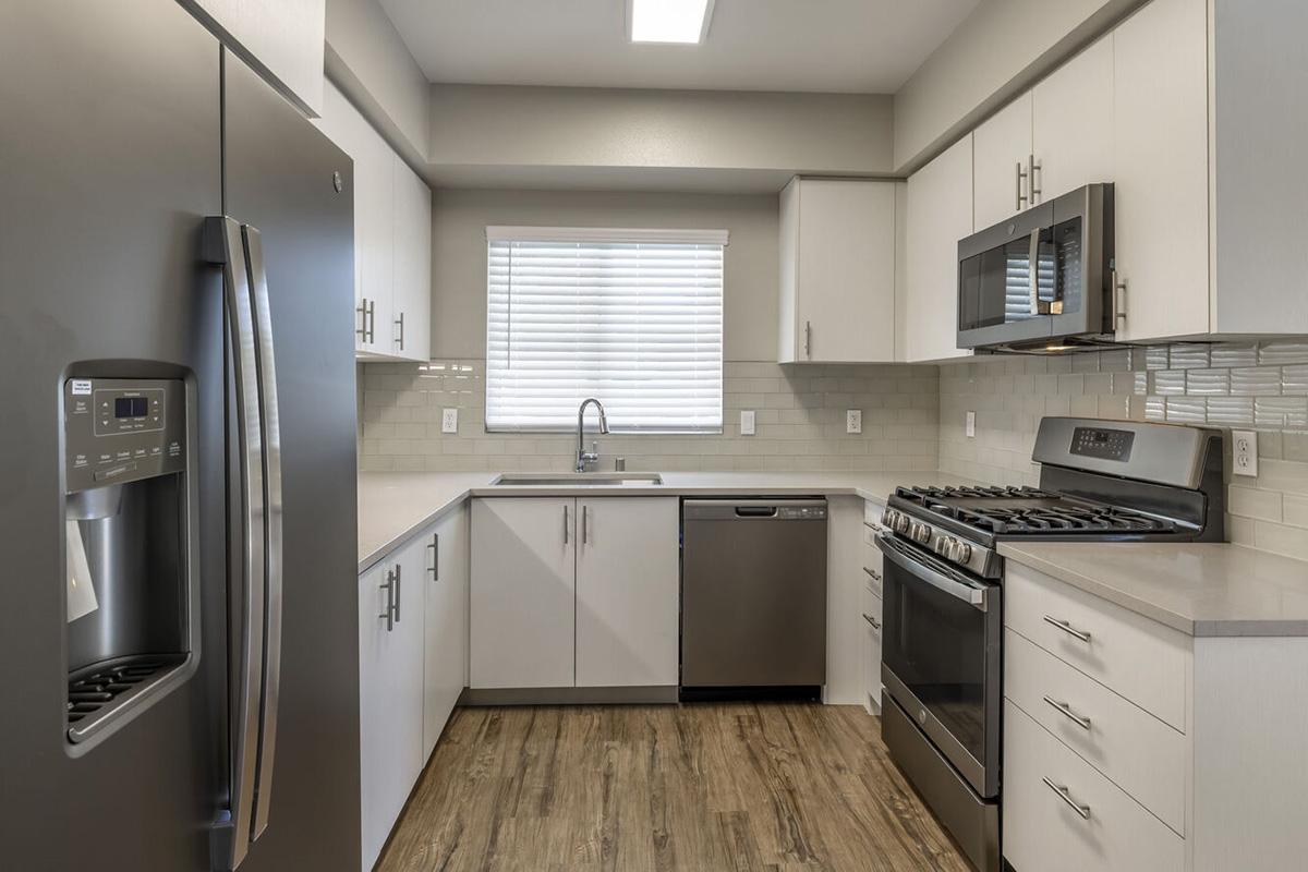 Modern kitchen with stainless steel appliances, including a refrigerator, microwave, and gas stove. Features white cabinetry, a gray countertop, and a dishwasher. Brightly lit with an overhead light and window providing natural light. Wood-style flooring complements the contemporary design.
