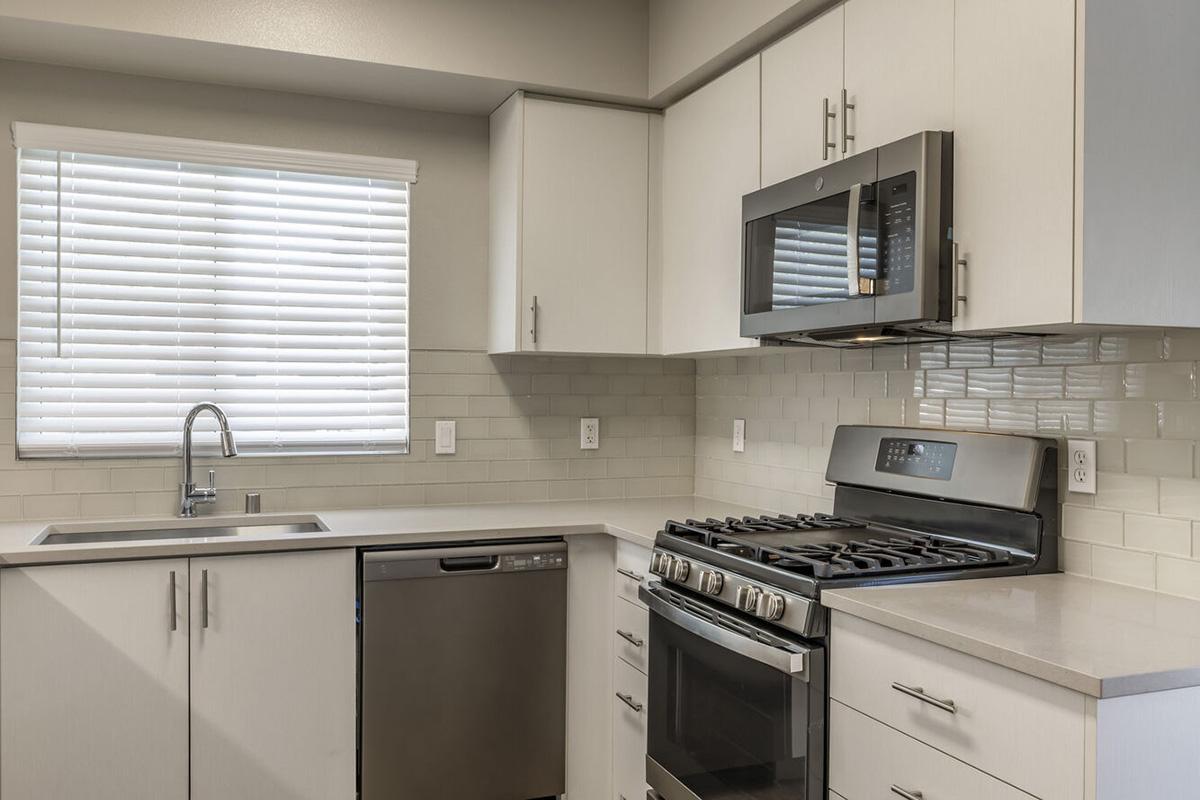 Modern kitchen featuring light-colored cabinetry, stainless steel appliances including a microwave, gas stove, and dishwasher. A large window with white blinds provides natural light, and the countertops are a light stone surface, complemented by a tiled backsplash.
