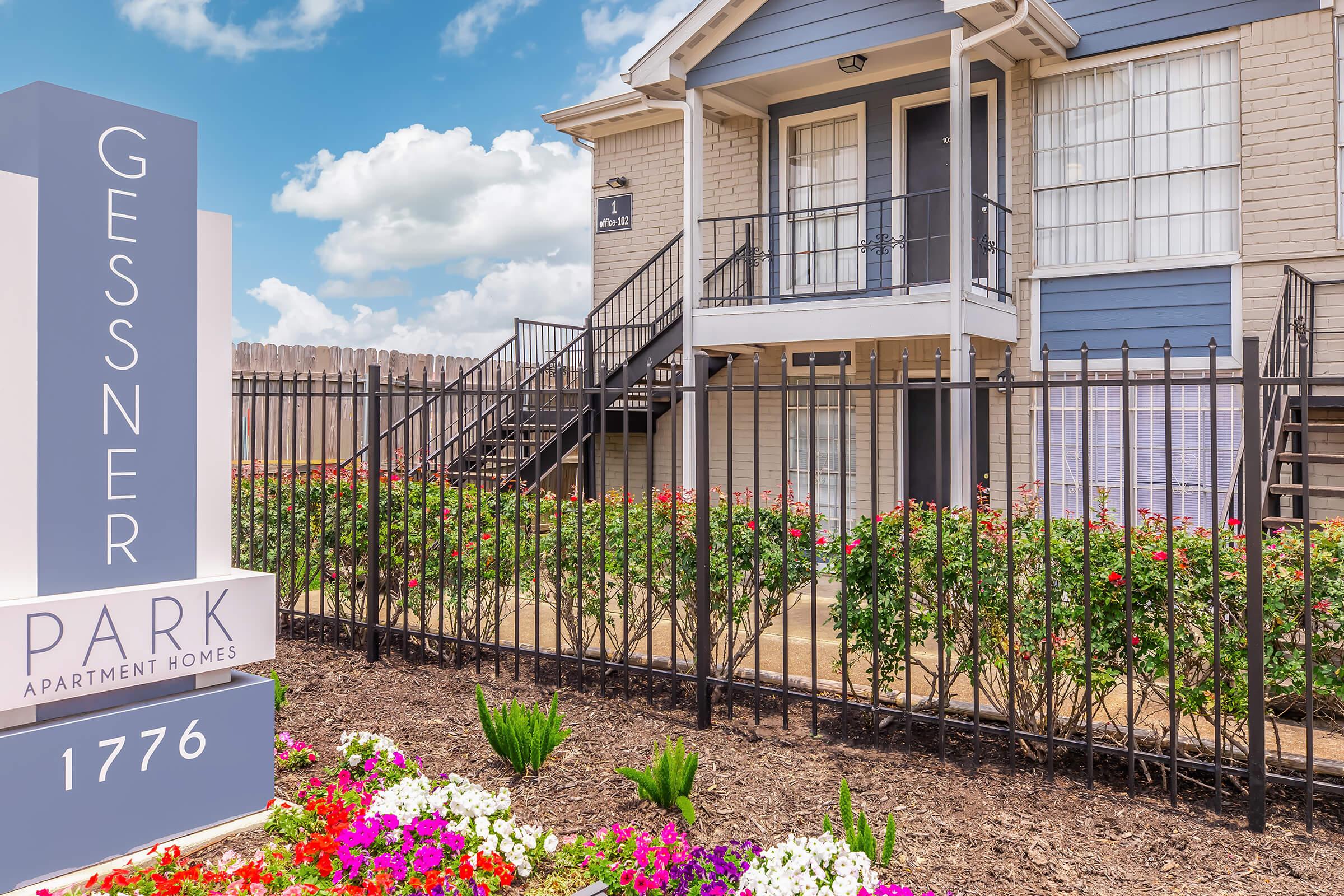 Exterior view of Gessner Park Apartment Homes, featuring a multi-story building with blue and beige siding. There are stairs leading up to the entrance, surrounded by a well-kept garden with colorful flowers. A sign indicating the property name and number is visible in the foreground. Bright blue sky with clouds in the background.