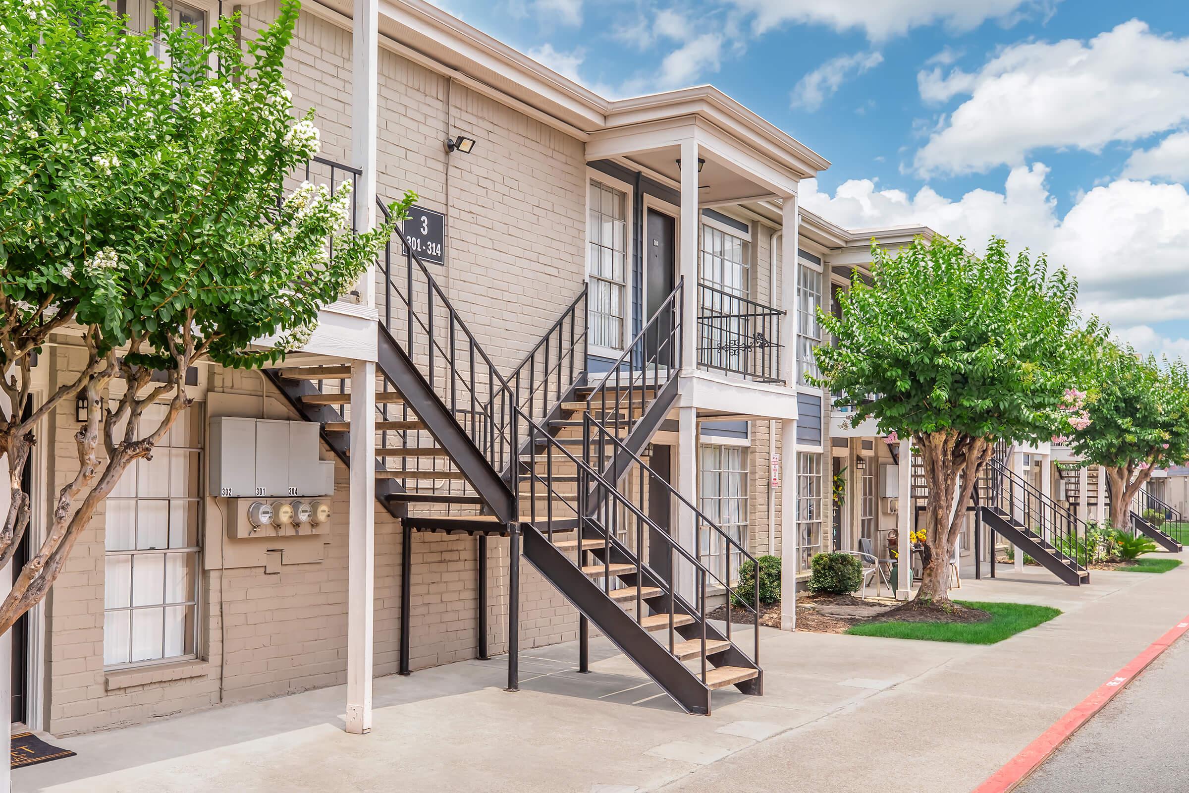 Row of outer apartment units featuring staircases leading to upper floors. Lush, green trees border the walkway, with shrubs and utility meters visible. The sky is bright with scattered clouds, creating a welcoming atmosphere.