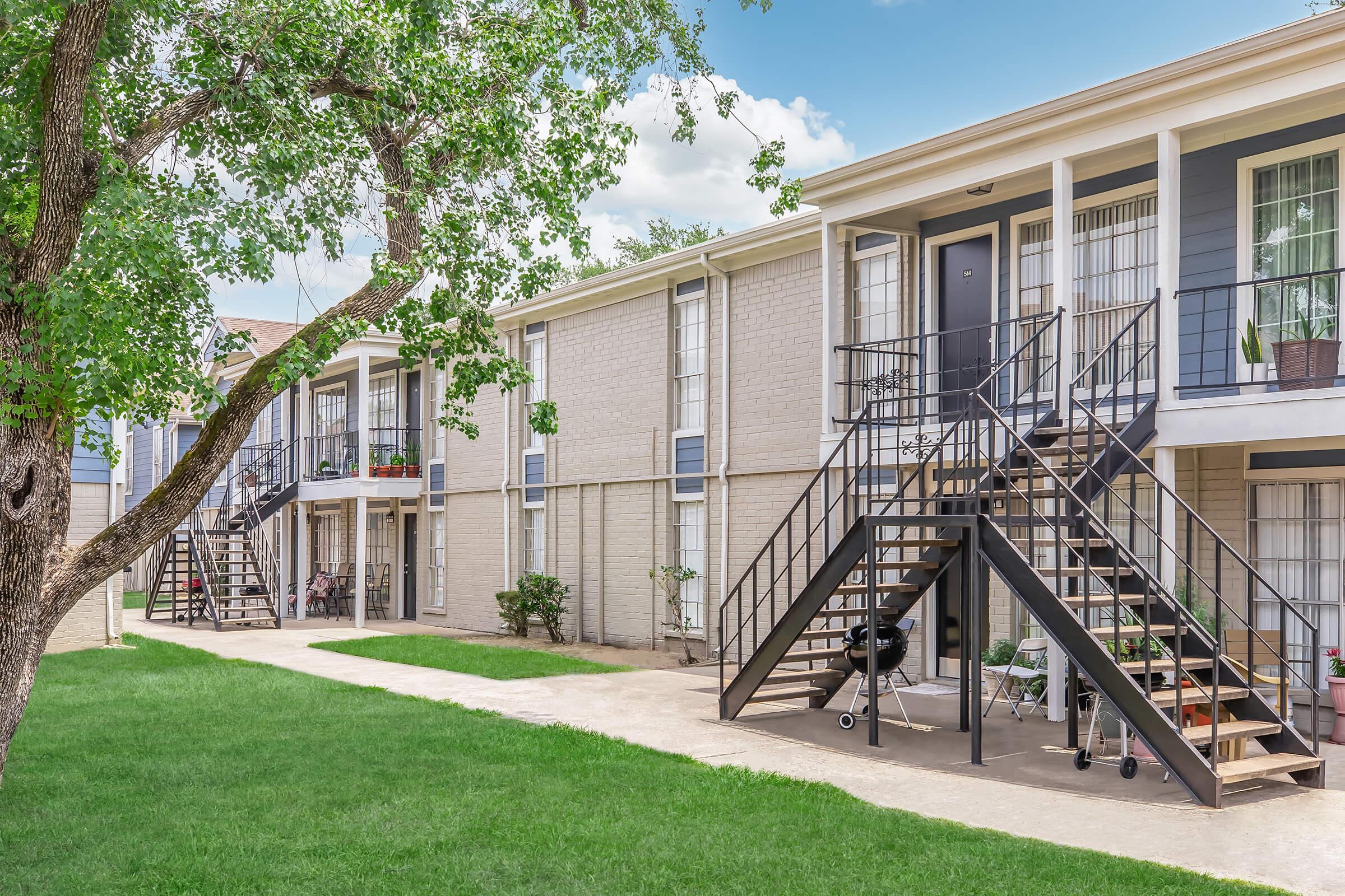 A landscaped courtyard with green grass and a large tree. In the background, two-story apartment buildings featuring gray and beige exteriors, with metal staircases leading to second-floor units. Potted plants are visible on some balconies, under a bright blue sky.