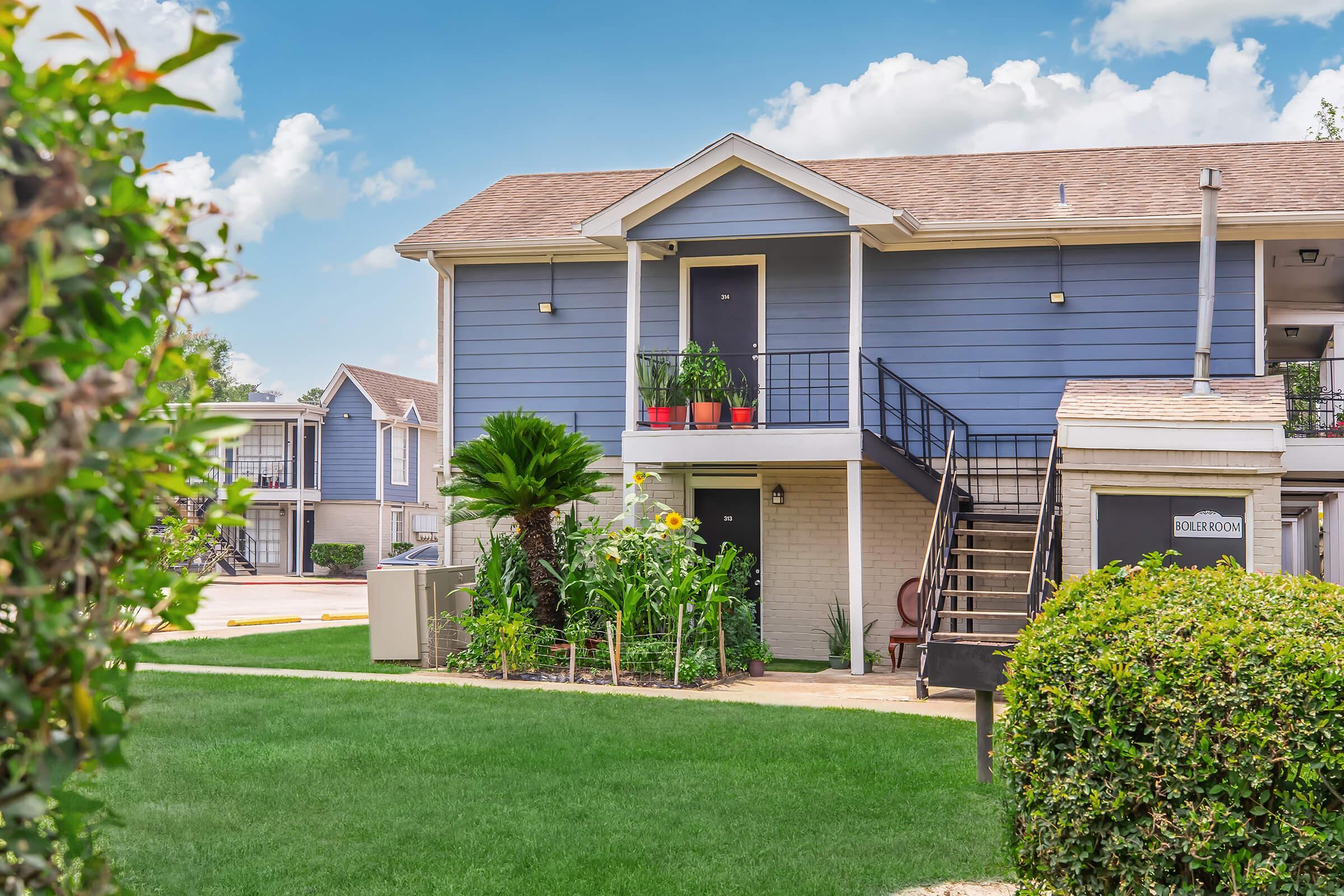 A landscaped exterior view of an apartment building featuring a two-story structure with a blue exterior, stairs leading to an upper door, and potted plants. Lush green grass surrounds the building, with a hedge and palm trees in the foreground. Bright blue sky with fluffy clouds completes the scene.