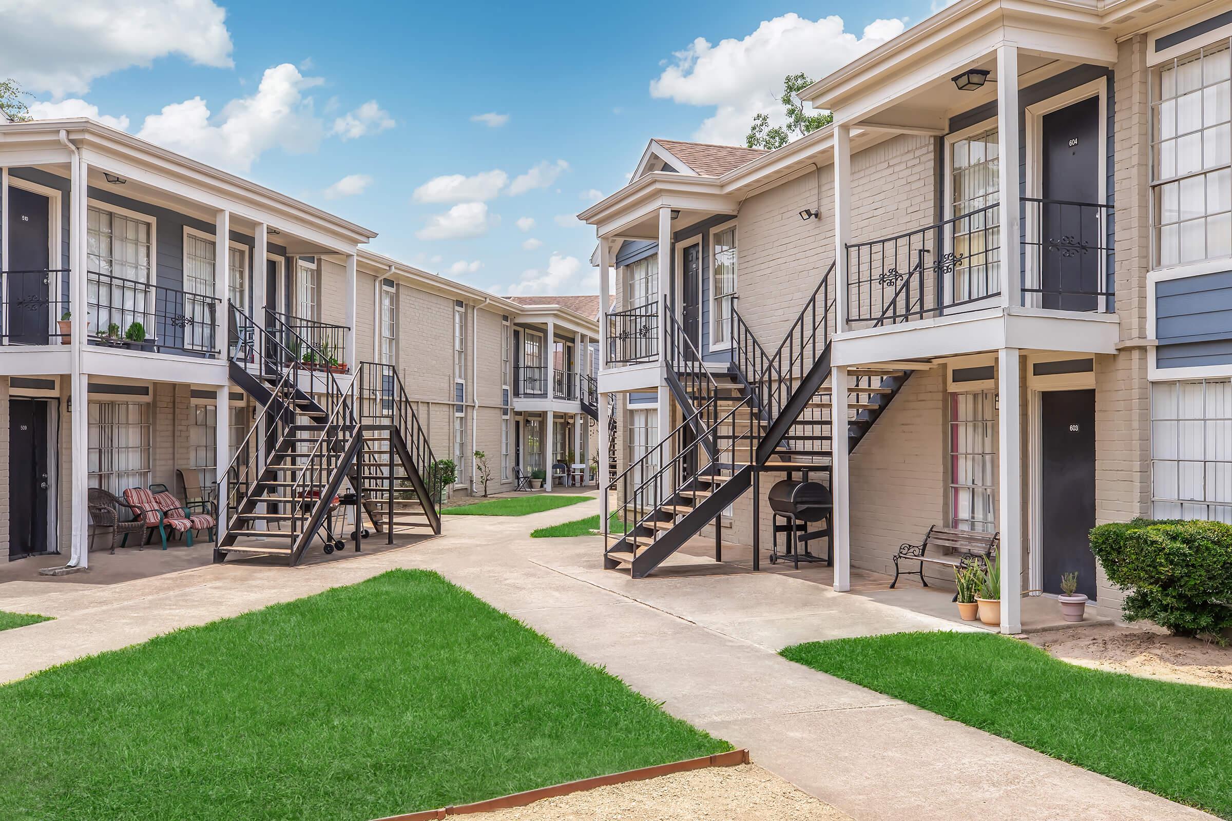 A view of a courtyard surrounded by two-story apartment buildings, featuring staircases leading to upper units. Lush green grass covers the ground, with a pathway running through the center. Some outdoor seating is visible, along with potted plants near the staircases. The sky is bright with scattered clouds.