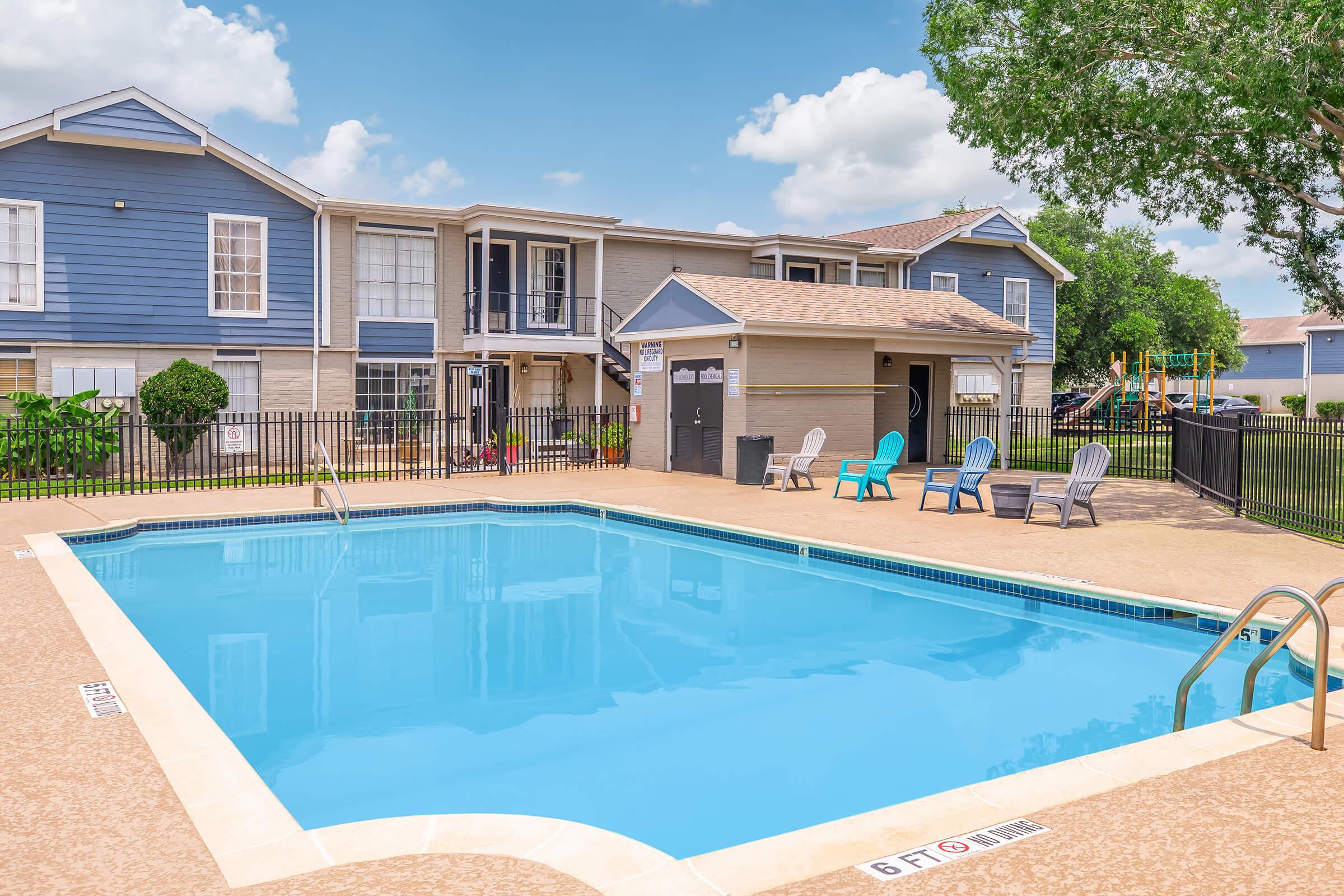 A clean, blue swimming pool surrounded by a tan deck, featuring several colorful lounge chairs. In the background, there are modern multi-story apartment buildings and a storage unit. Lush green trees and a bright blue sky complete the scene, creating a relaxing outdoor atmosphere.