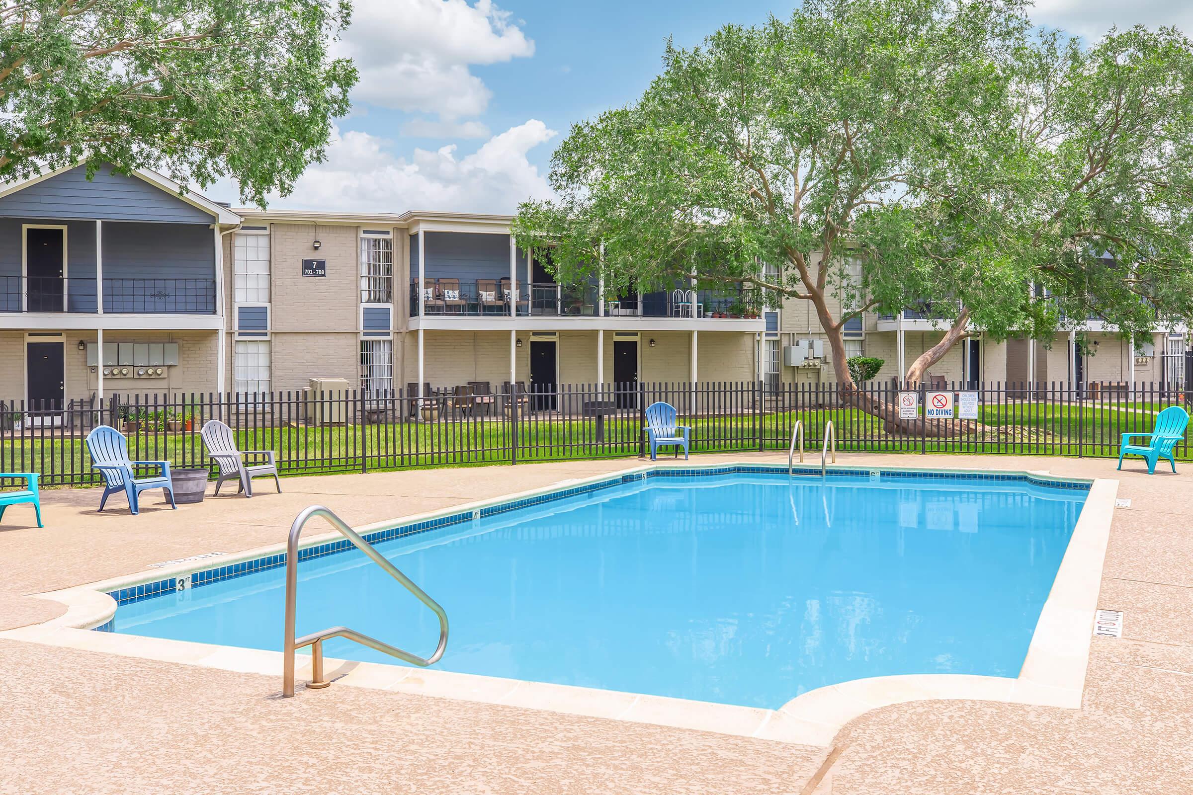 A clear blue swimming pool surrounded by a concrete deck. Lounge chairs are placed around the pool area. In the background, an apartment complex with multiple levels is visible, featuring balconies and a well-maintained lawn. A fence encloses the pool area, providing privacy. Bright, sunny day with a few clouds in the sky.