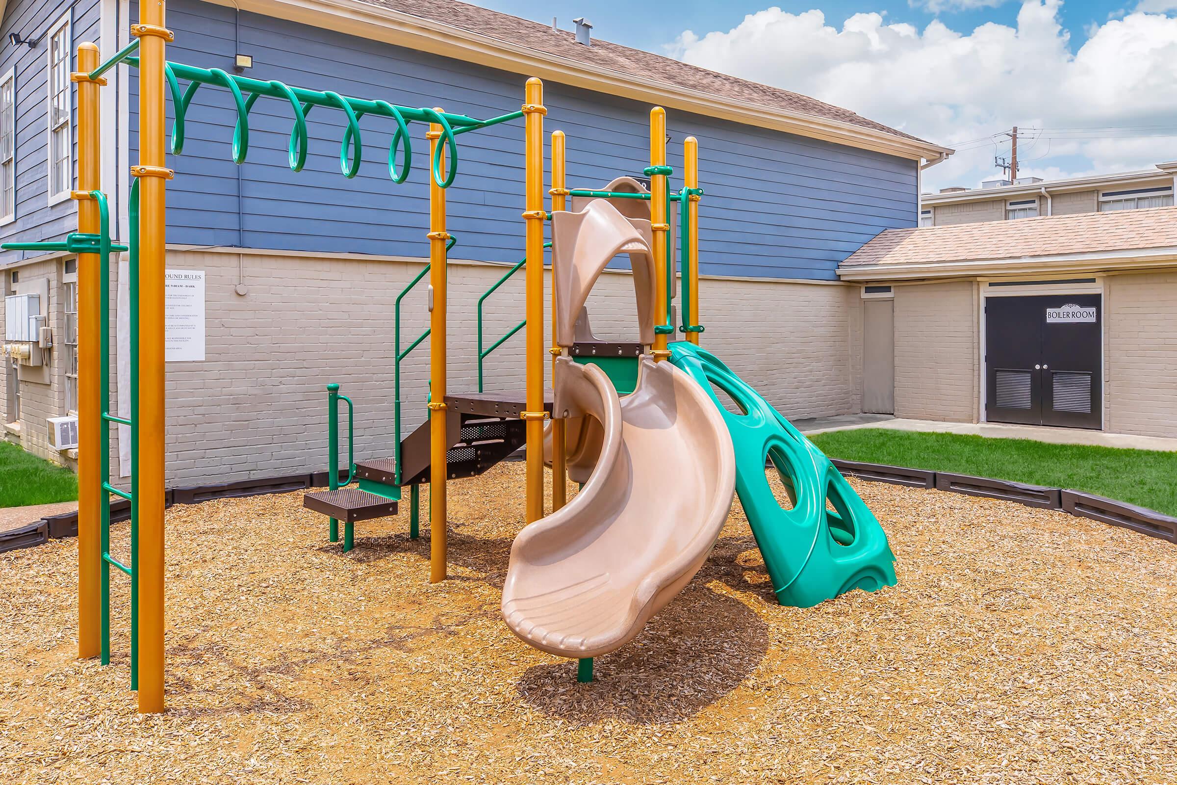 A playground featuring a brown slide and a green climbing structure. The playground is surrounded by tan gravel and is located next to a light-colored building with blue siding. The sky above is bright with scattered clouds.