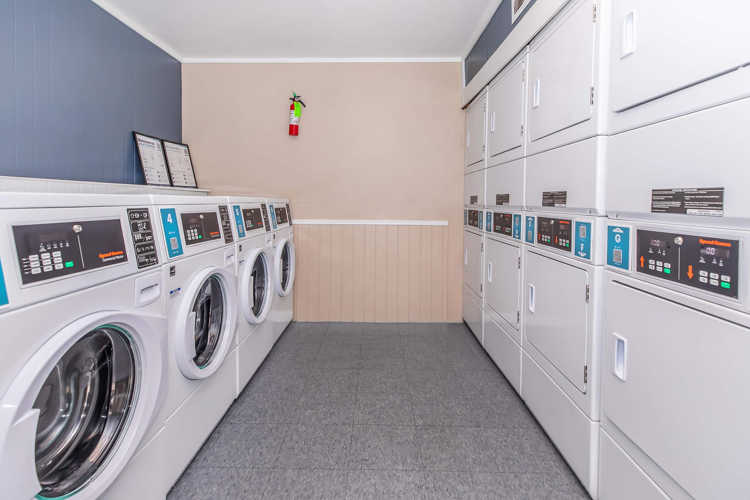 A clean, well-organized laundromat featuring multiple rows of modern white washing machines and dryers. The walls are painted a light tan with a blue accent, and a small decorative item is visible on the wall. The floor is tiled, and there are instruction signs and payment machines visible.