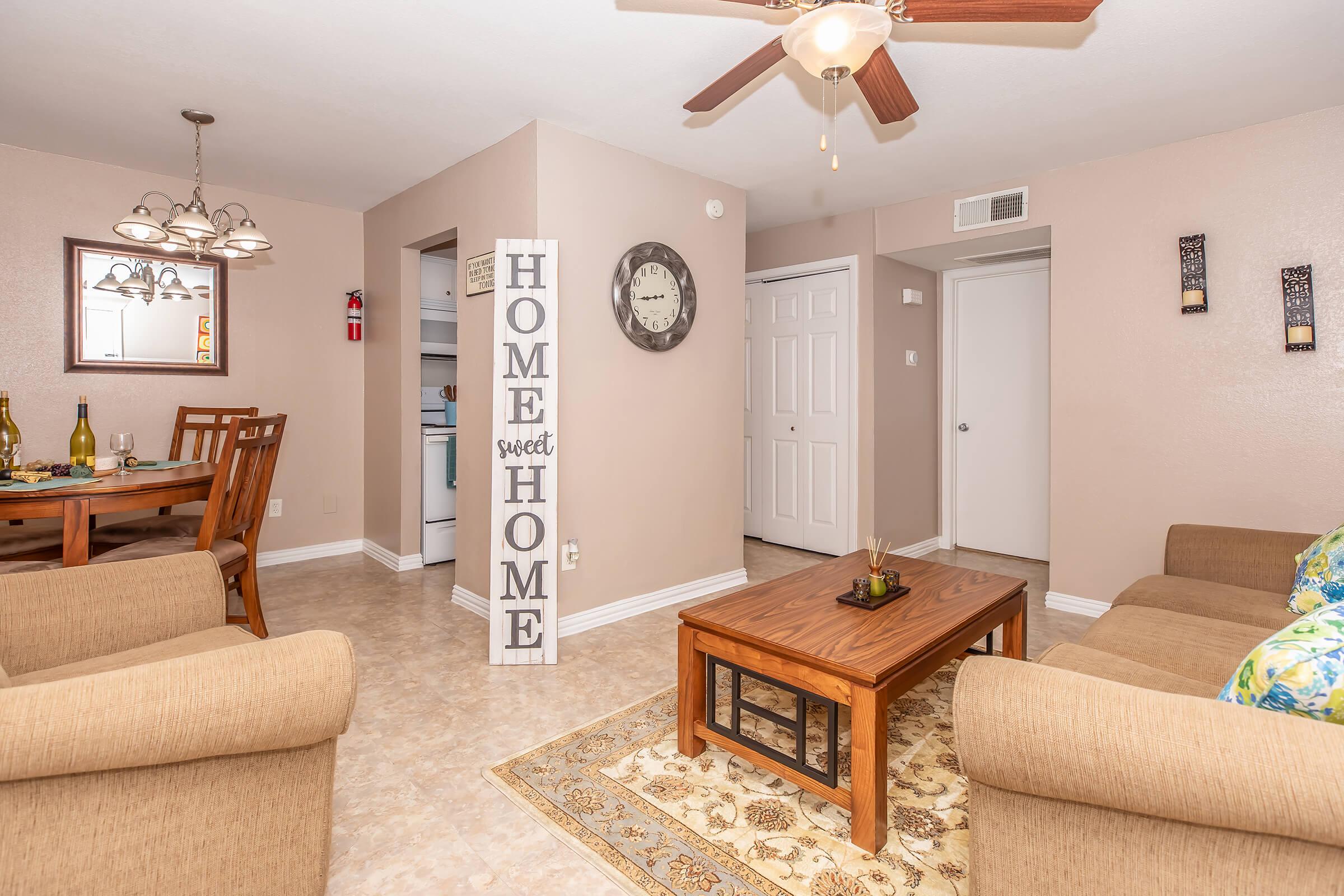 A cozy living space featuring a beige couch, a wooden coffee table, and decorative elements. A wall sign reads "HOME sweet HOME." The dining area includes a wooden table with chairs, and there is a wall clock next to an entrance. The decor is warm and inviting, with neutral tones and patterned accents.