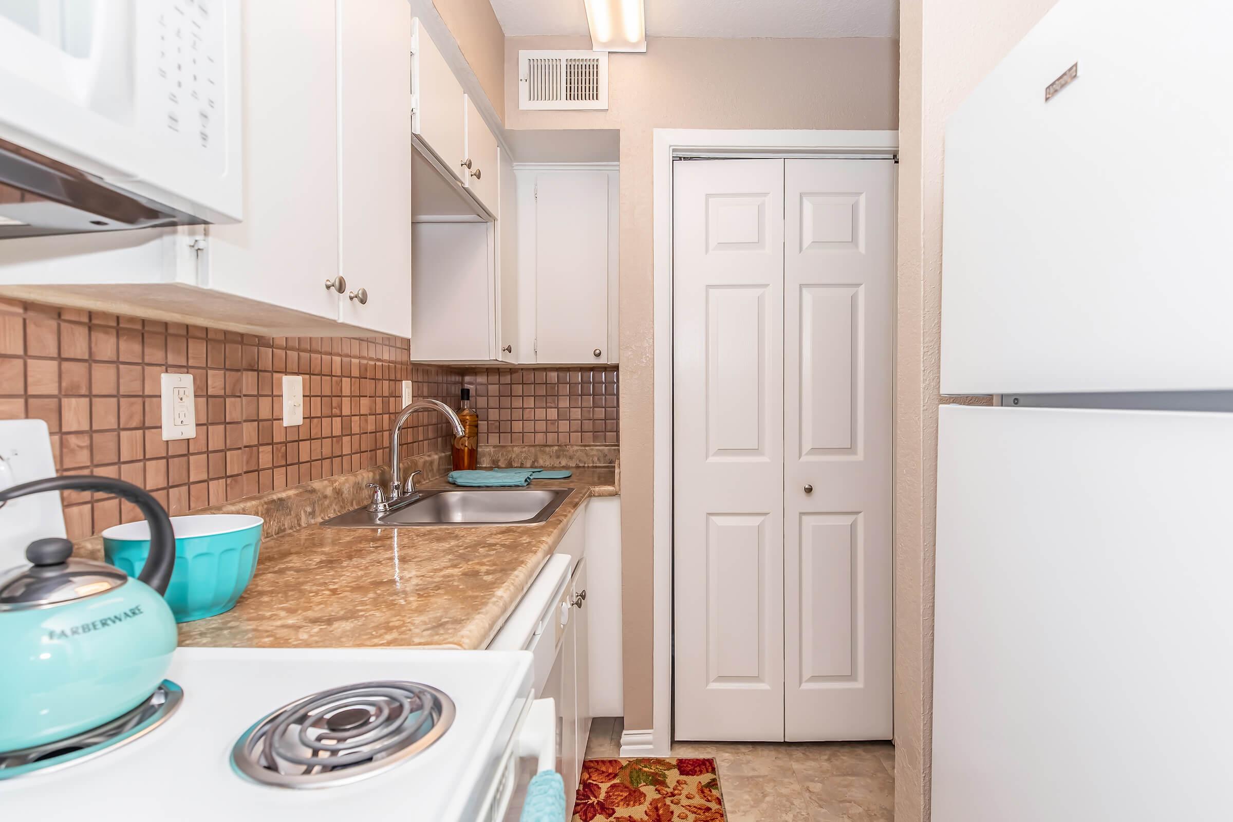 A kitchenette featuring white cabinets, a light-colored countertop, and a stainless steel sink. There is a stove, a microwave, and a refrigerator. The backsplash is made of brown tiles, and the floor is tiled. A rug adds a pop of color, and a turquoise bowl and kettle are visible on the countertop.