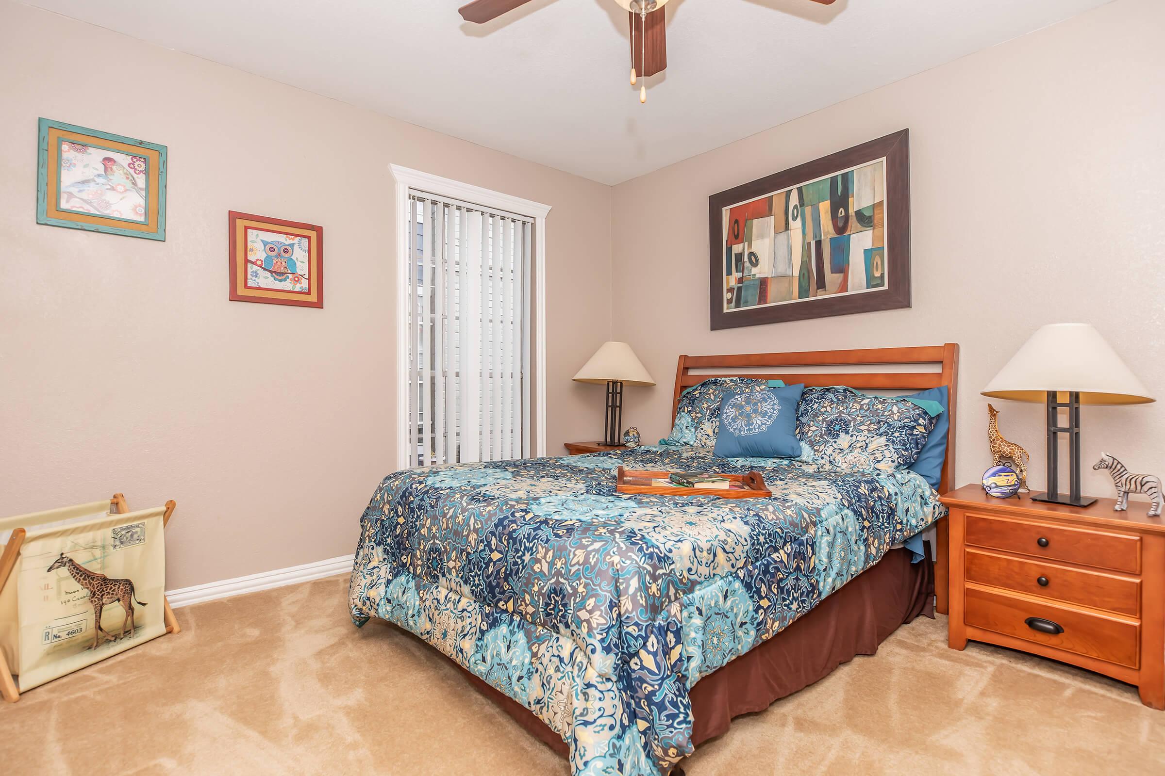 A cozy bedroom featuring a queen-sized bed with a colorful blue and brown patterned quilt. There are bedside lamps on wooden nightstands, decorative artwork on the walls, and a window with sheer curtains. The room has beige walls and carpet, creating a warm and inviting atmosphere.