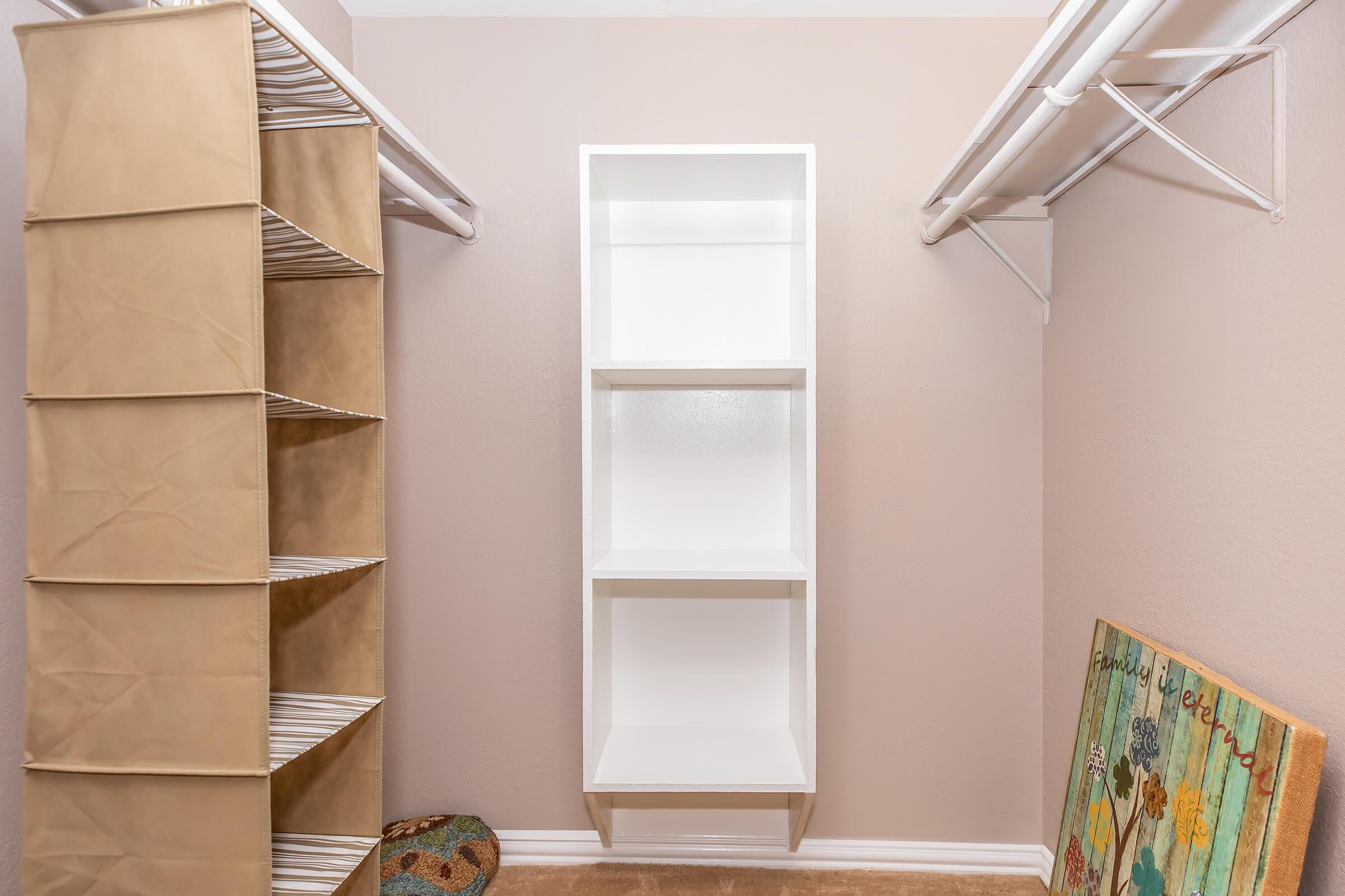 A neatly organized closet featuring a beige fabric shelving unit on the left and a white open shelving unit on the right. The walls are painted light brown, and there's a colorful wall art piece and a round decorative object on the carpeted floor. The space is well-lit and looks tidy.