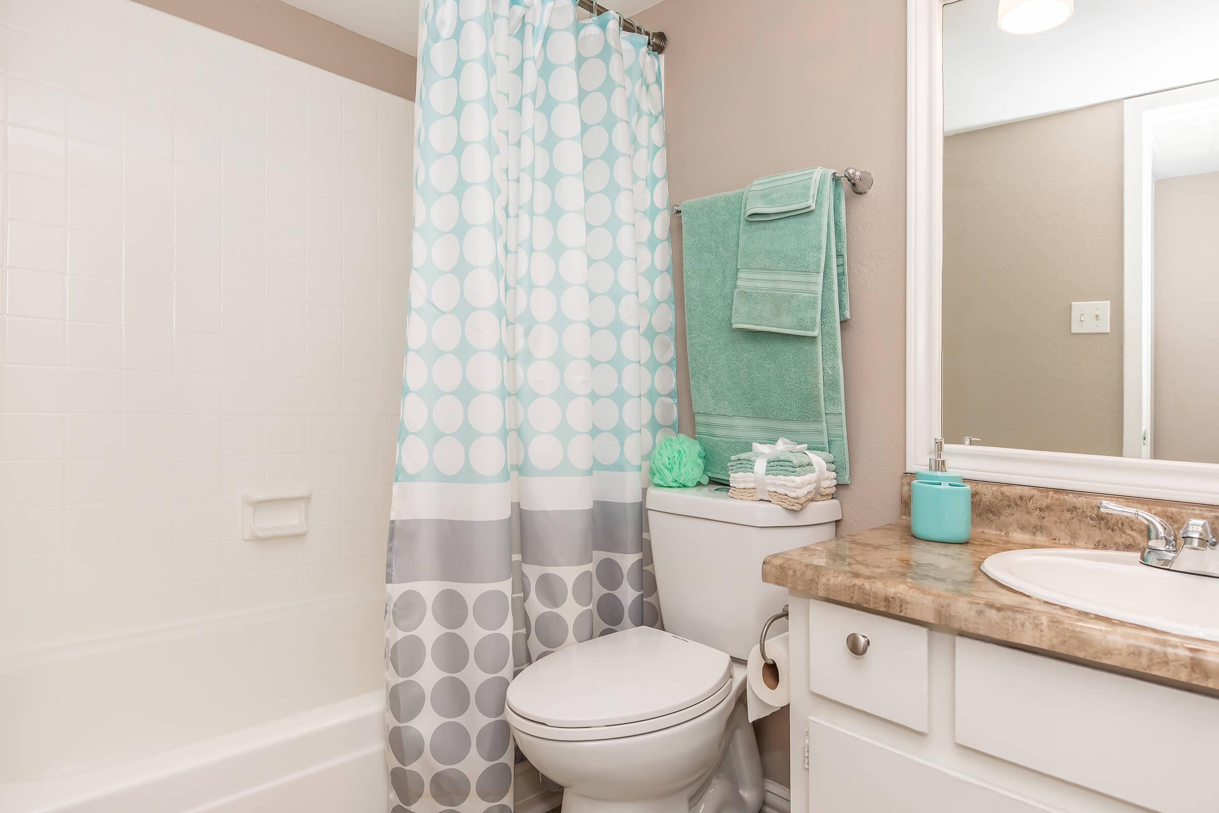 A clean and modern bathroom featuring a white bathtub with a light blue and gray polka dot shower curtain. There's a toilet beside the tub, decorated with a teal towel and bath accessories on the counter. A mirror above the sink reflects the neutral-colored walls and ample lighting.
