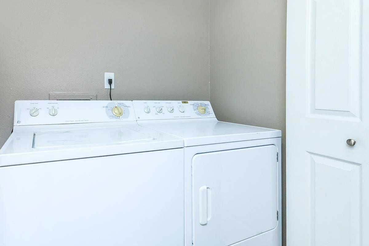 A white washing machine and dryer stacked next to a closed white door against a light brown wall. The laundry appliances are equipped with control knobs and dials for operation. The space appears clean and well-maintained.