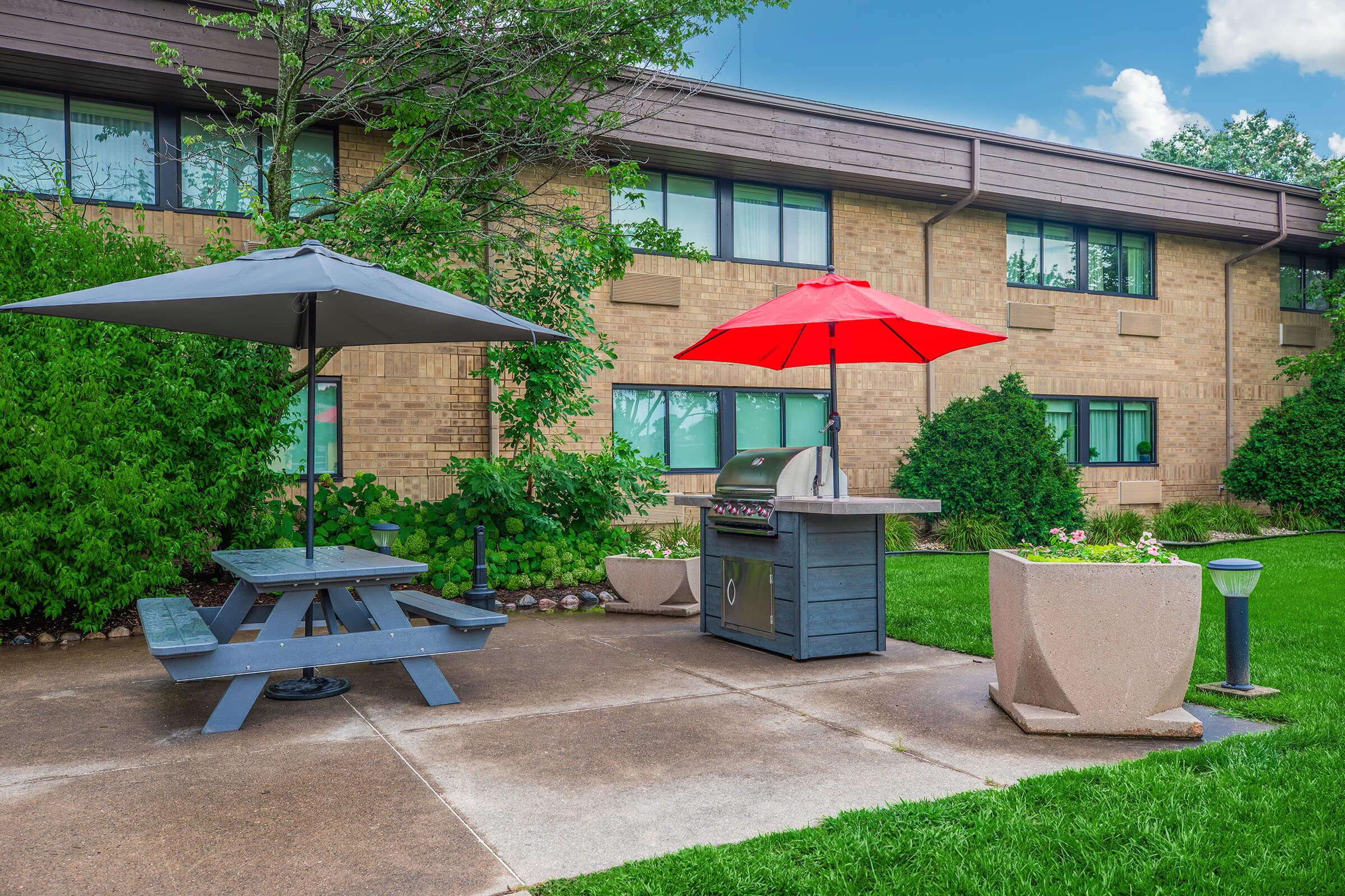 A backyard patio area featuring a charcoal grill under a gray umbrella, with a red umbrella providing shade over a picnic table. The setting includes a decorative planter and lush greenery in the background, alongside a brick building with windows. The sky is partly cloudy, indicating a pleasant outdoor atmosphere.