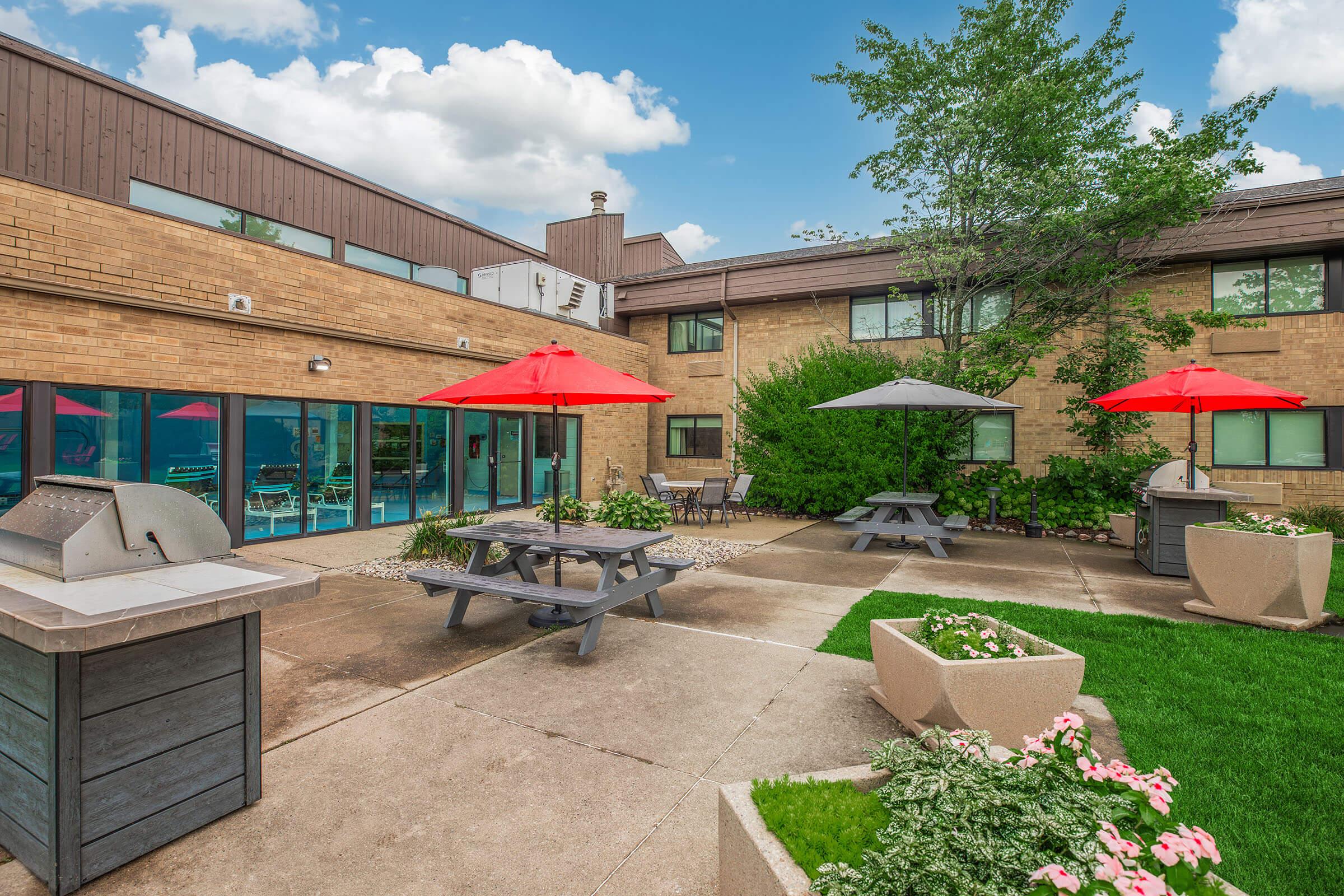 A spacious outdoor area featuring picnic tables with red and gray umbrellas, surrounded by greenery and flower beds. In the background, there is a building with large windows, and a barbecue grill is visible. The sky is blue with scattered clouds, creating a pleasant atmosphere.