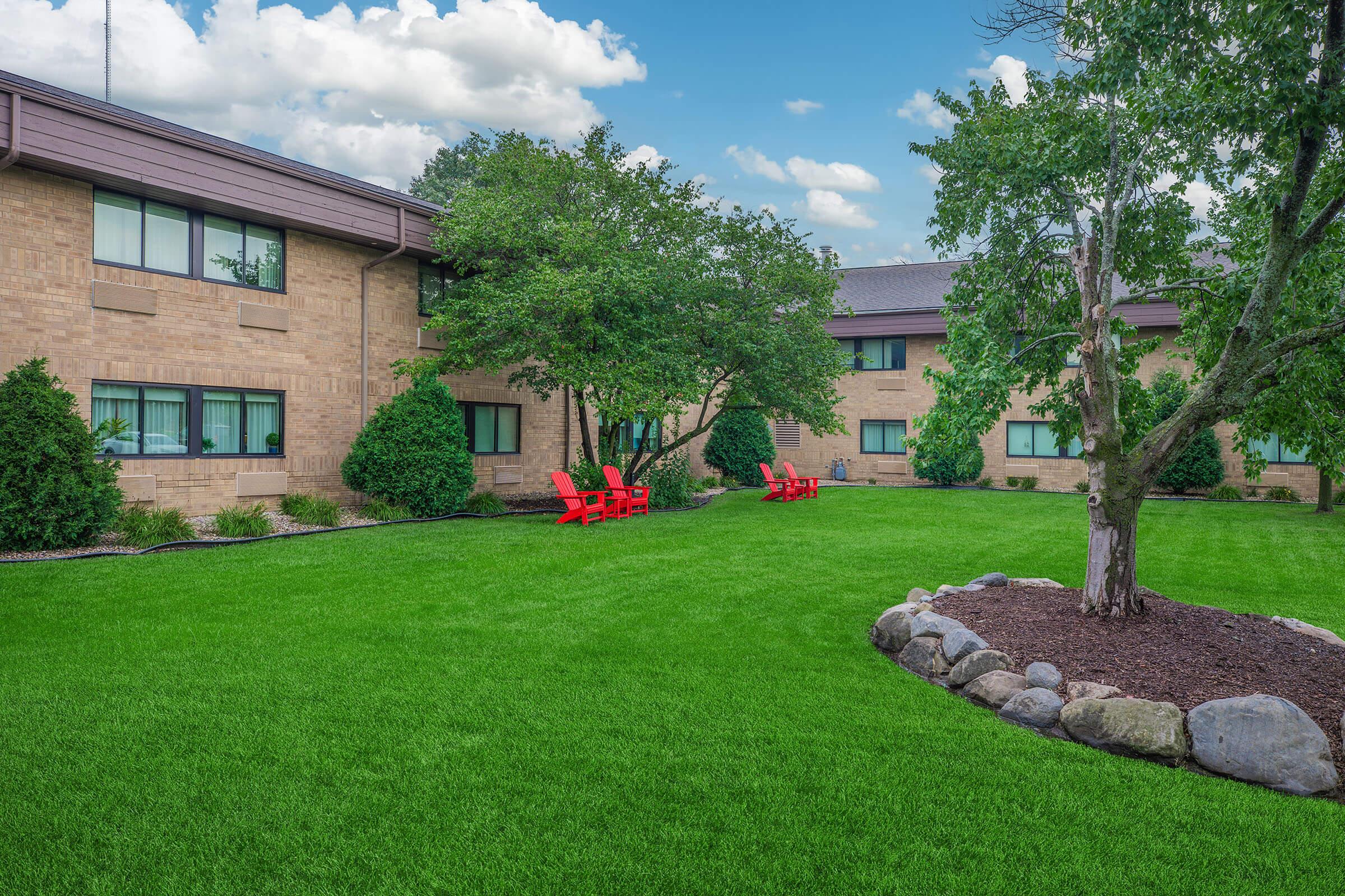 A well-maintained lawn with vibrant green grass, featuring red Adirondack chairs arranged in a cozy seating area. Surrounding the chairs are lush shrubs and a small tree, with a backdrop of a brown brick building under a blue sky with scattered clouds. A decorative rock border edges the garden area.