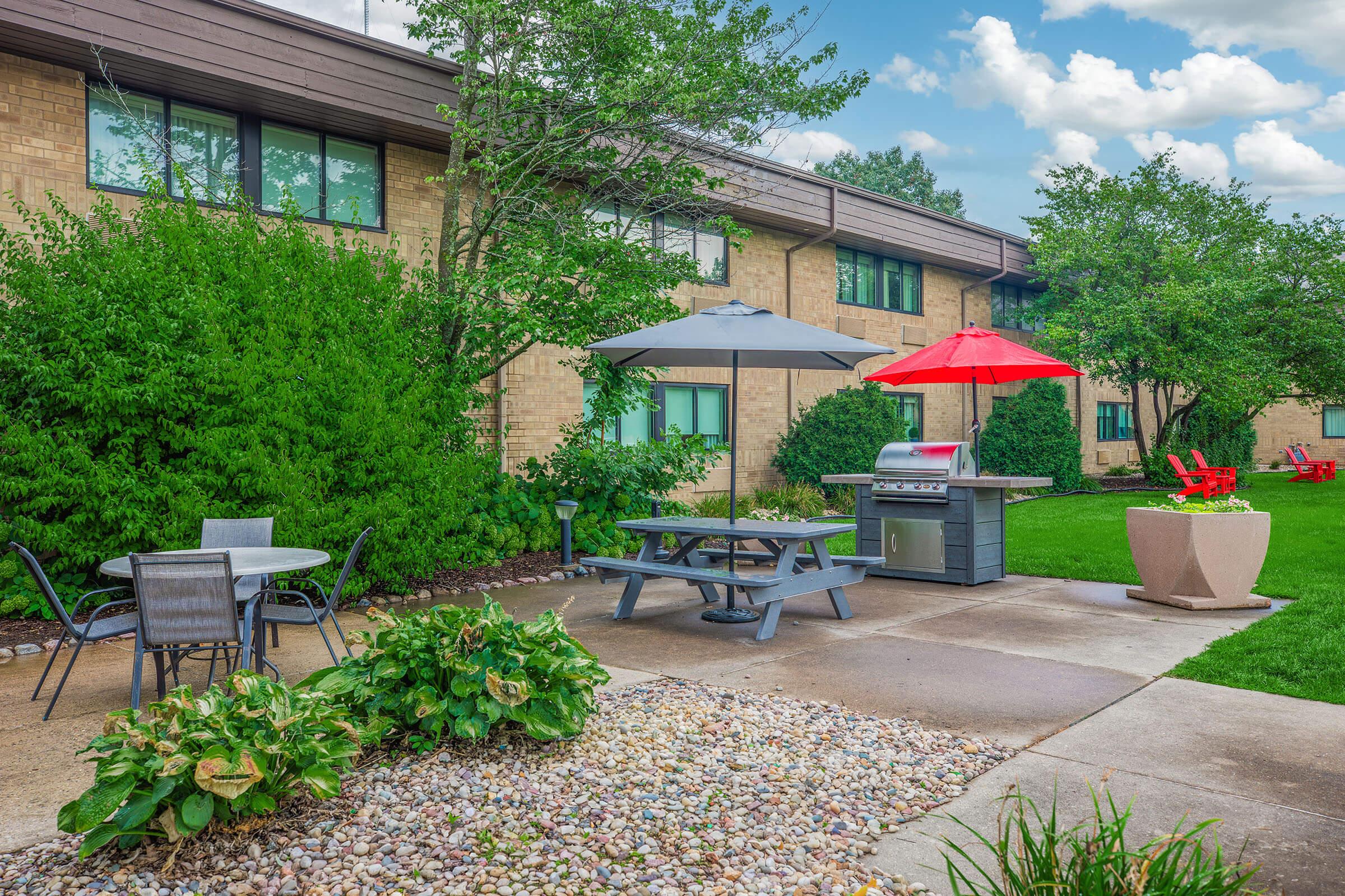 A landscaped outdoor area featuring a picnic table with a grill, surrounded by green bushes and trees. There are two patio umbrellas—one gray and one red—and a seating arrangement with chairs. The building in the background has multiple windows and a grass lawn.