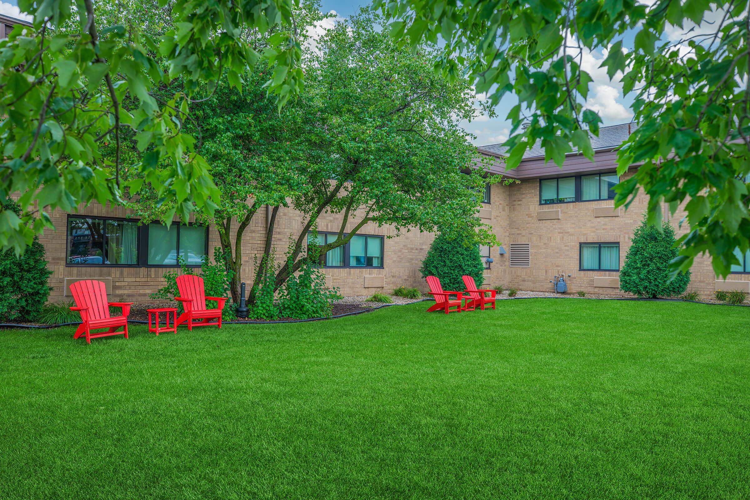 A peaceful outdoor area featuring a lush green lawn bordered by trees. In the foreground, bright red Adirondack chairs and a small table create a cozy seating arrangement, inviting relaxation. The background shows a stone building with large windows and well-maintained shrubs.