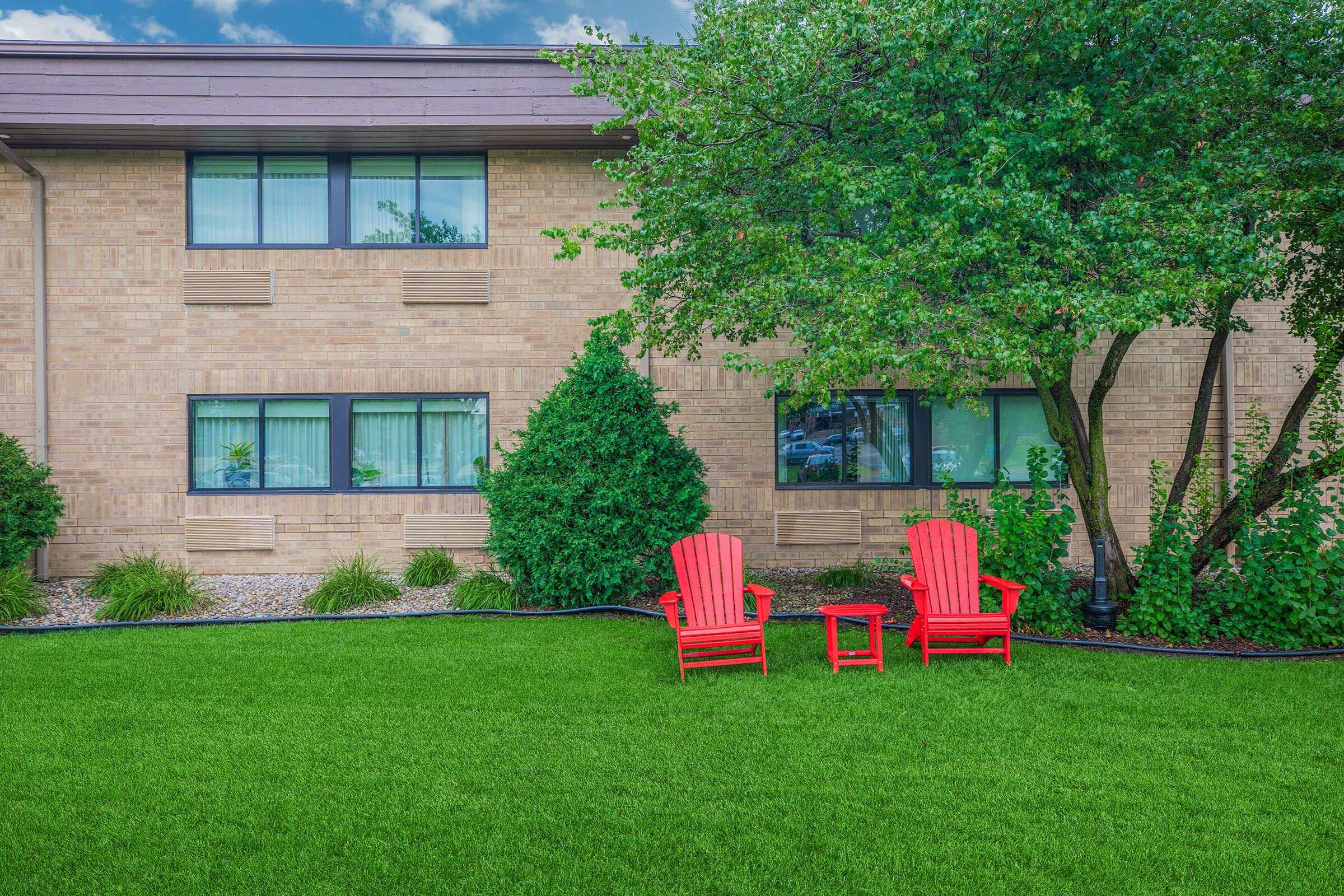 A vibrant green lawn features two red Adirondack chairs and a small side table, set against a backdrop of a brick building with large windows and greenery. A tree provides shade, enhancing the cozy outdoor space, inviting relaxation and enjoyment.