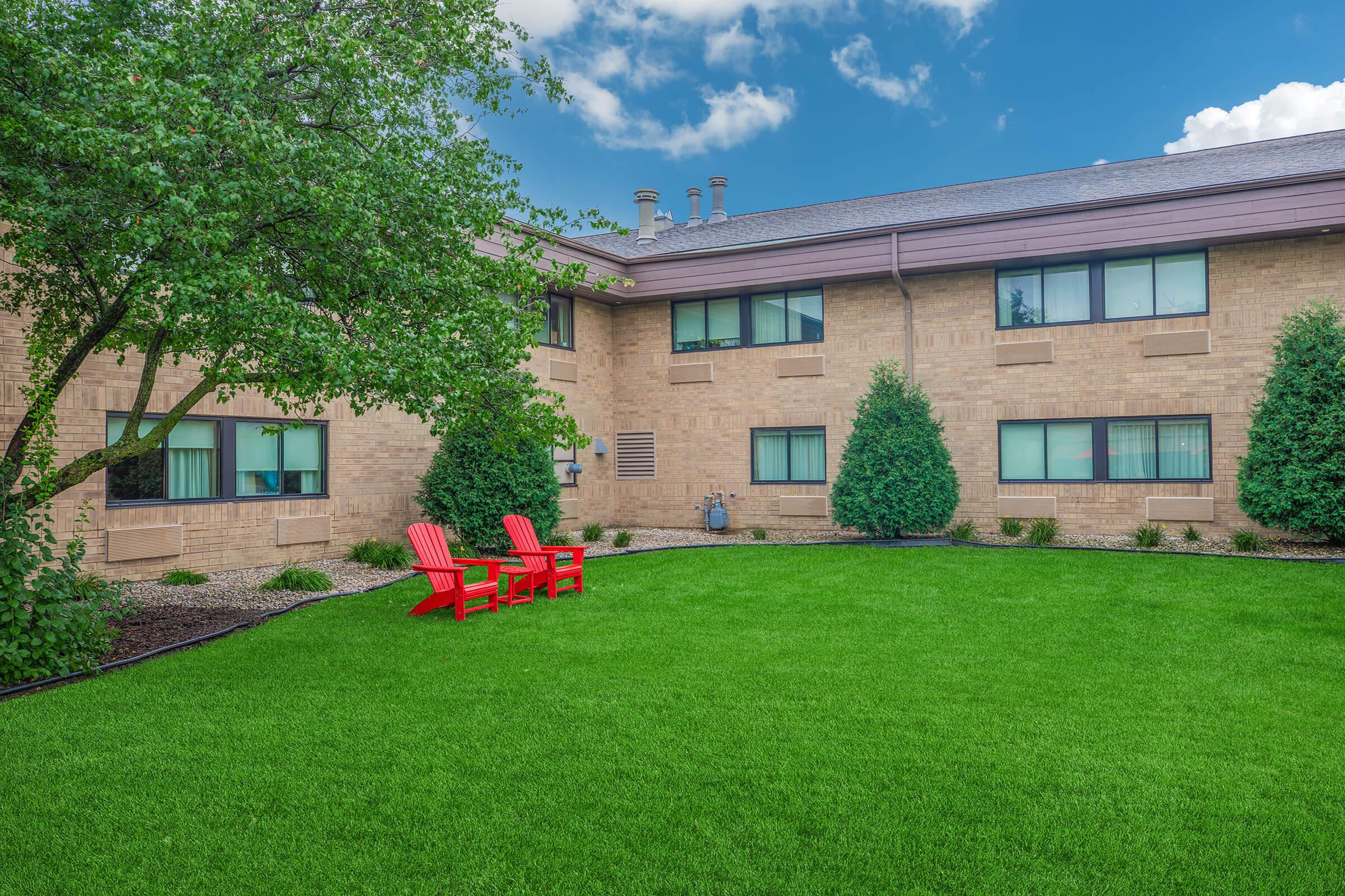 Green lawn area with two red Adirondack chairs positioned on the grass, surrounded by neatly trimmed bushes and a brick building with several windows. A tree provides additional greenery, and the sky features some clouds.