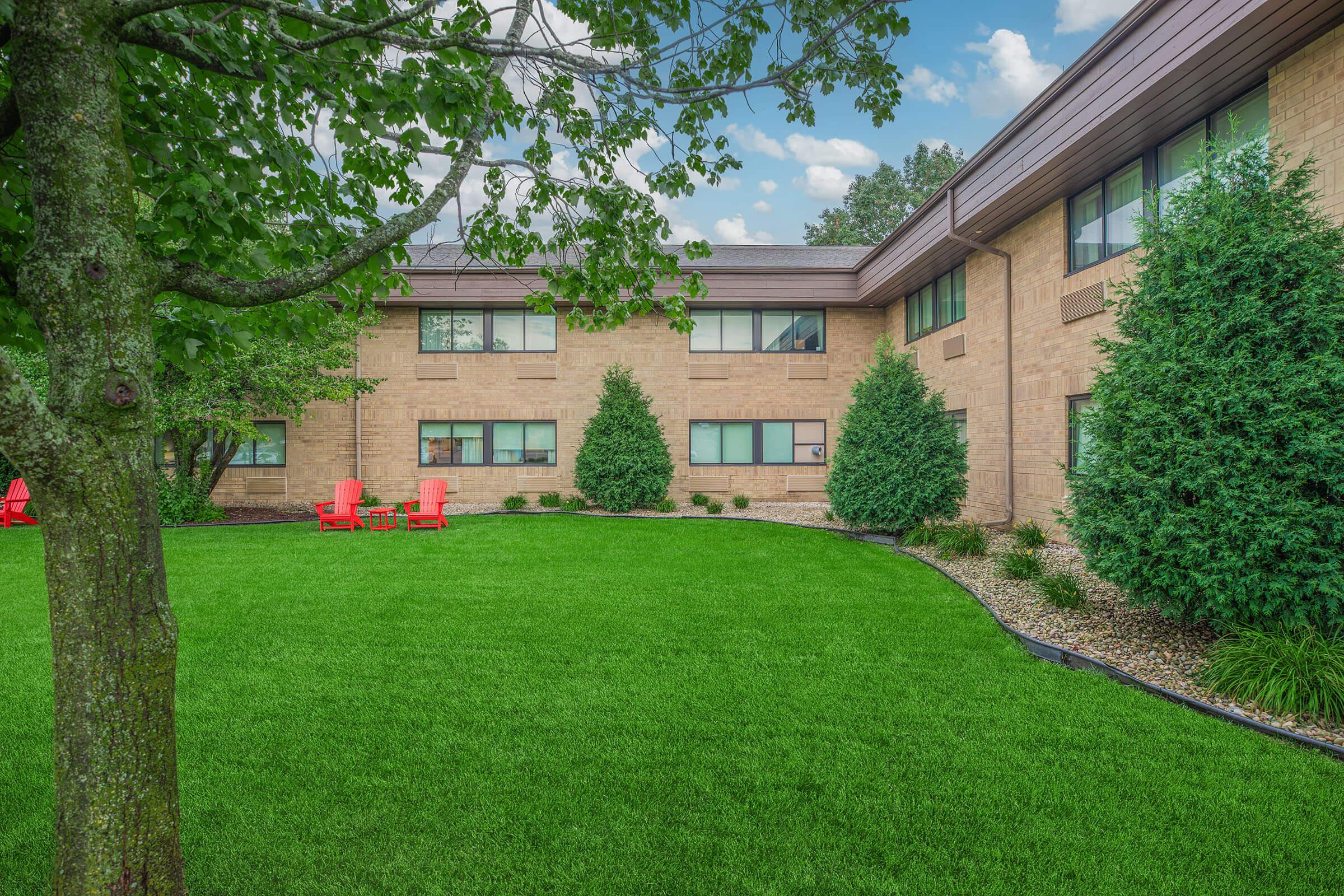 A well-maintained grassy area with two bright red outdoor chairs visible. The background features a brick building with several windows and lush greenery, including neatly trimmed bushes and trees. The sky is partly cloudy, adding a pleasant ambiance to the scene.