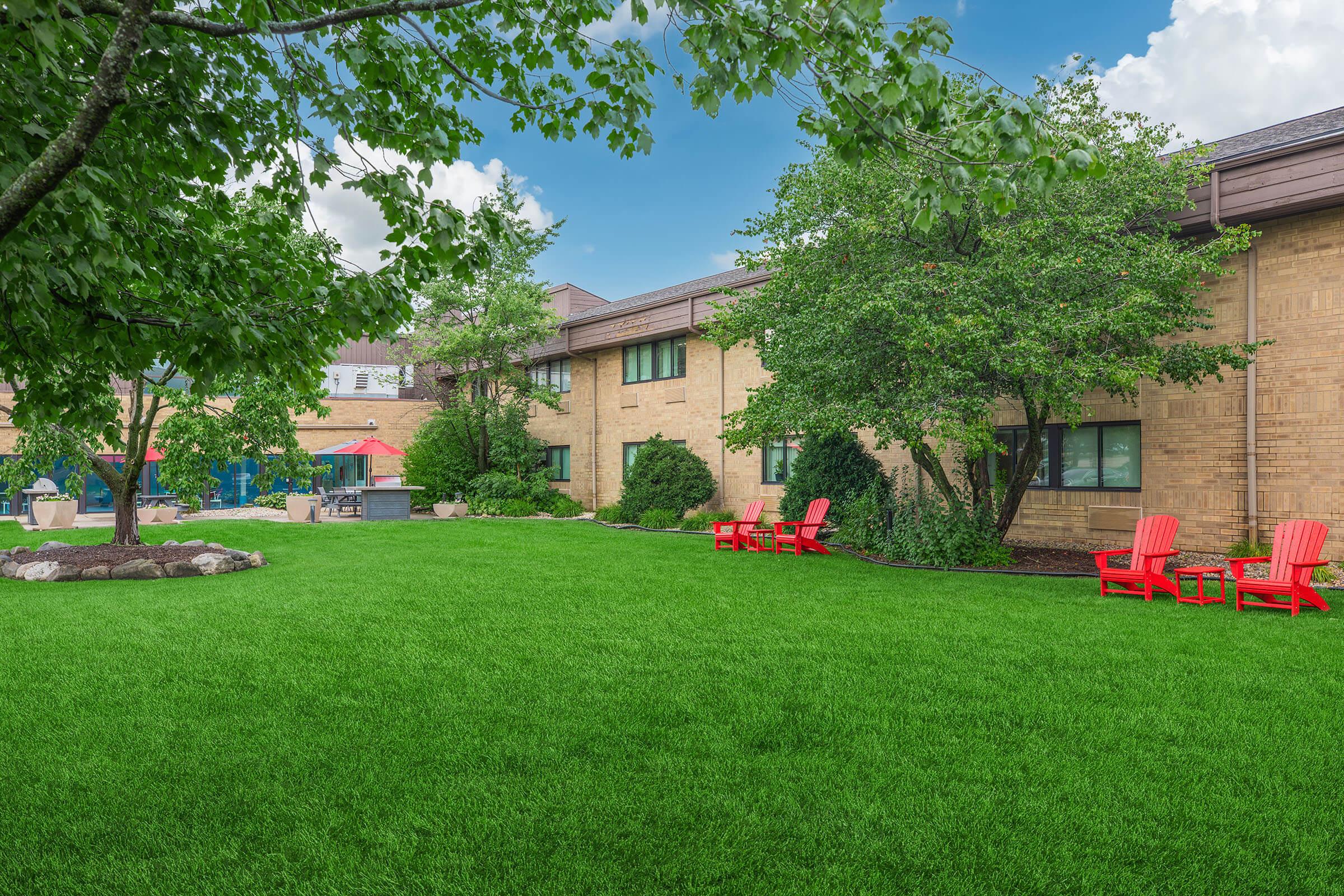 A well-maintained green lawn surrounded by trees, with bright red Adirondack chairs arranged in a casual seating area. In the background, a brick building is visible, along with a glimpse of outdoor amenities, creating a welcoming and serene atmosphere. A clear blue sky with clouds completes the scene.