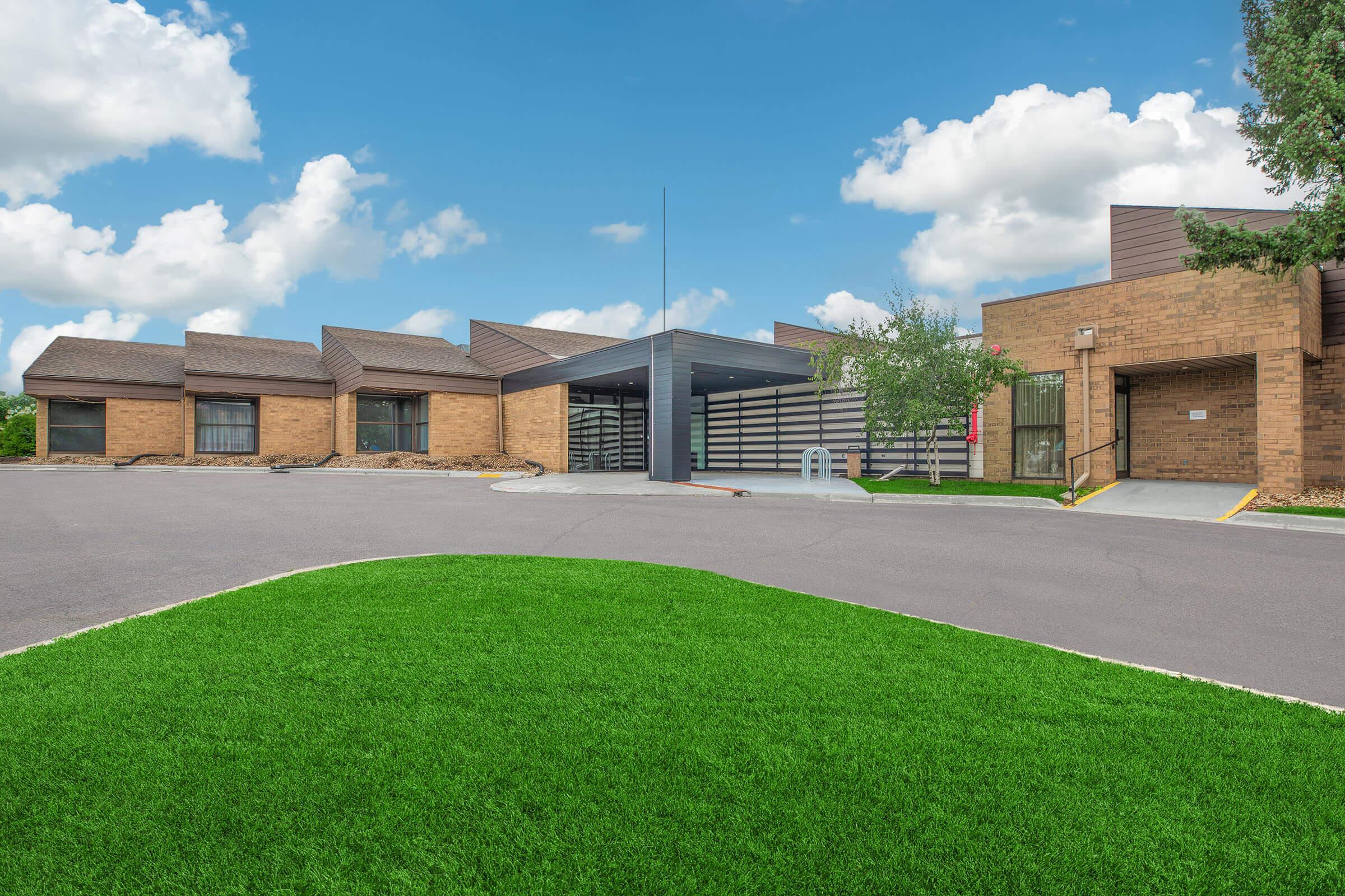 A modern building with a spacious entrance. The structure features a combination of brick and modern architectural elements, set against a clear blue sky with fluffy white clouds. Lush green grass lines the pathway leading to the entrance, creating a welcoming atmosphere.