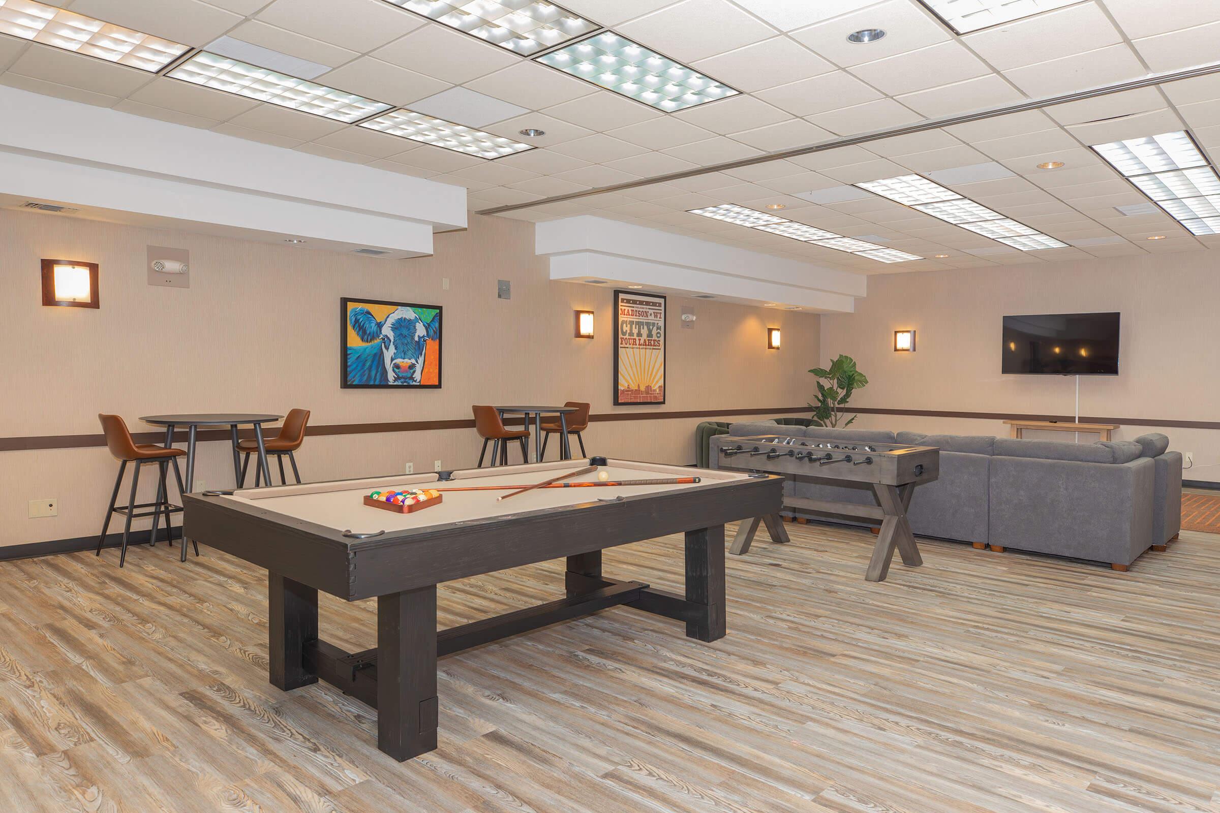 A modern recreational room featuring a pool table, foosball table, and comfortable seating. The space has a warm ambiance with stylish lighting and wooden flooring. A colorful cow painting decorates the wall, and there are tables and chairs for socializing. A TV is mounted on the wall, enhancing the entertainment options.