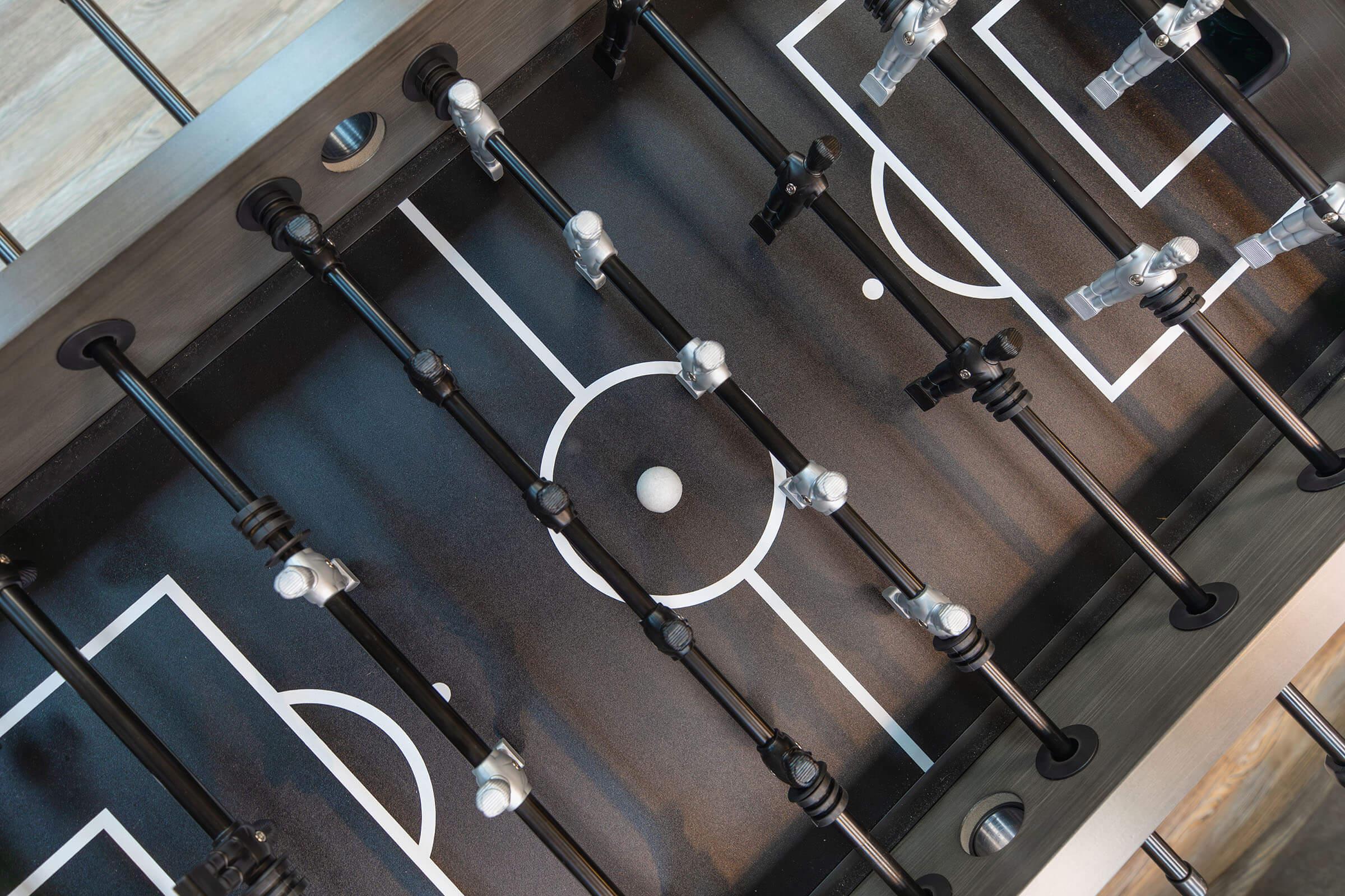 A top-down view of a foosball table, featuring black and silver player figurines arranged on a dark playing surface with white lines marking the field. A small white ball is positioned in the center of the table, ready for gameplay.