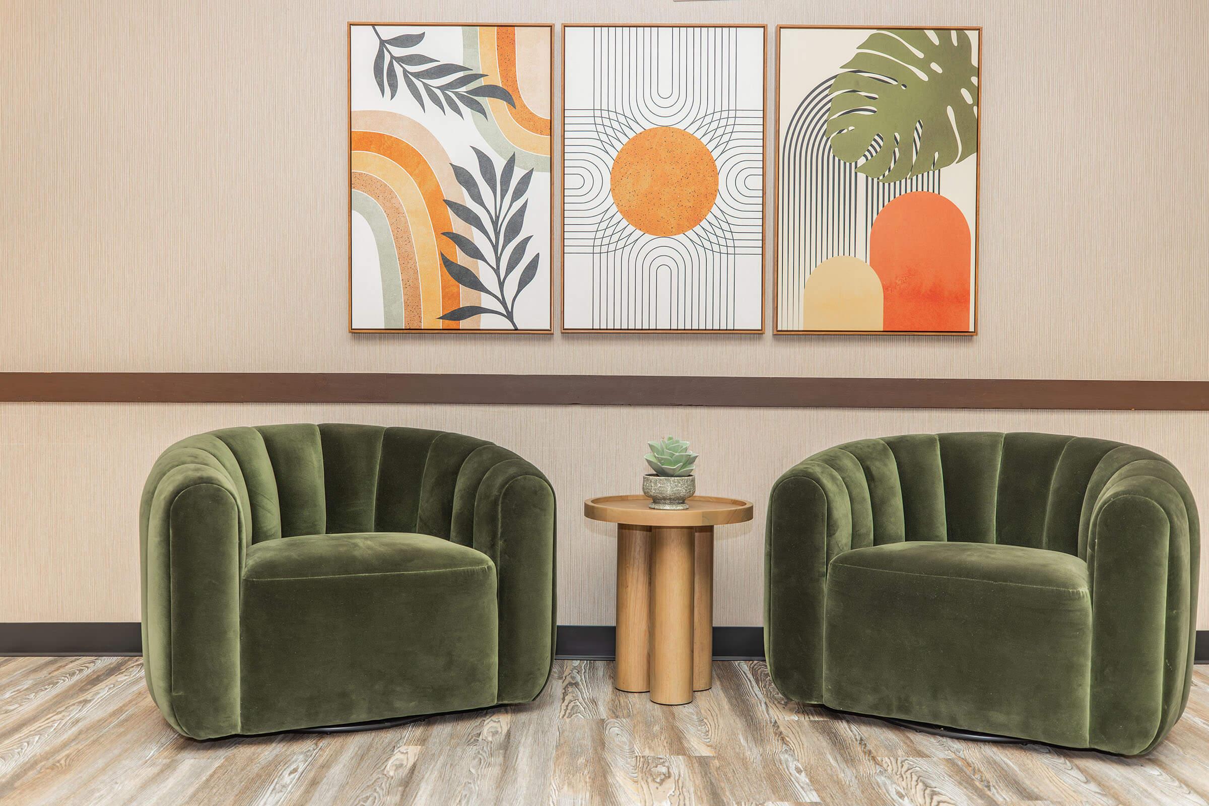 Two plush, green velvet armchairs are positioned facing each other beside a round wooden side table. The wall features three colorful paintings with abstract designs, including sun motifs and leafy patterns. A small succulent plant sits on the table, creating a cozy and inviting atmosphere.