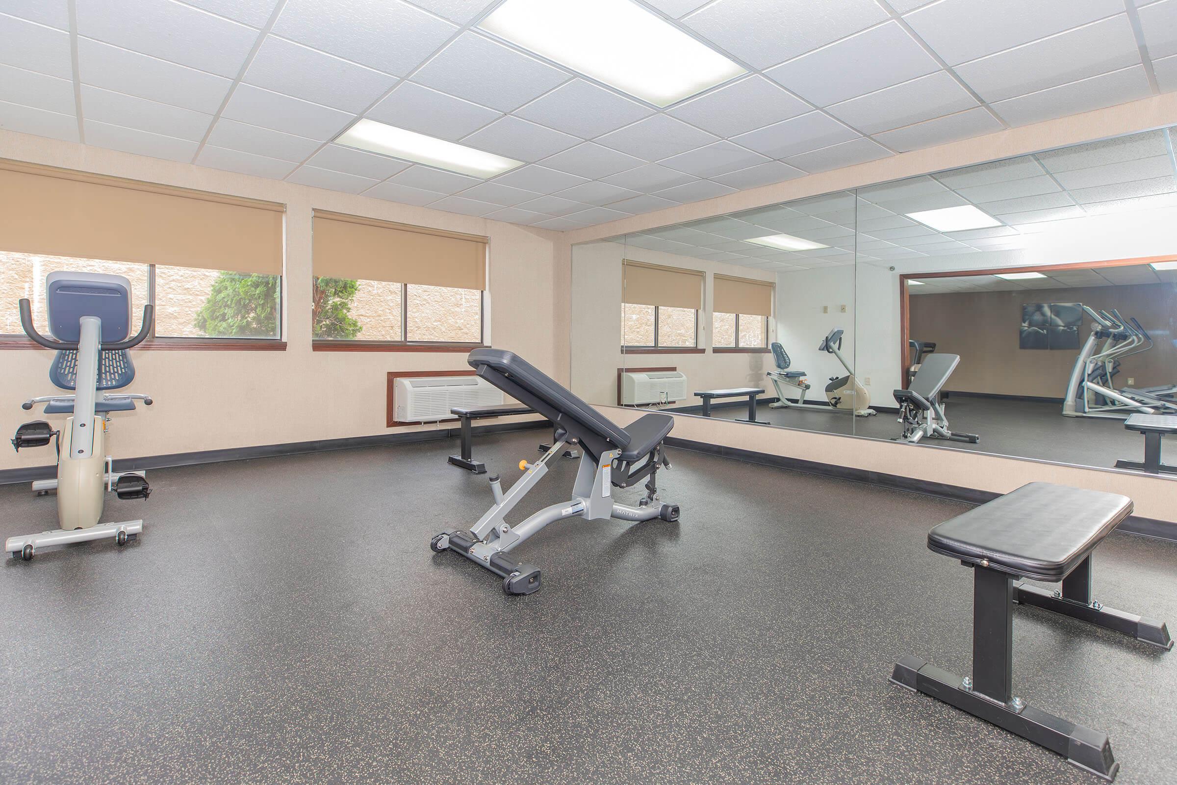 A modern gym interior featuring exercise equipment including a stationary bike, an adjustable bench, and a weight bench. The room has large mirrors on one wall and large windows with blinds, providing natural light and a clean, well-kept environment. The flooring is black rubber, enhancing the gym aesthetic.