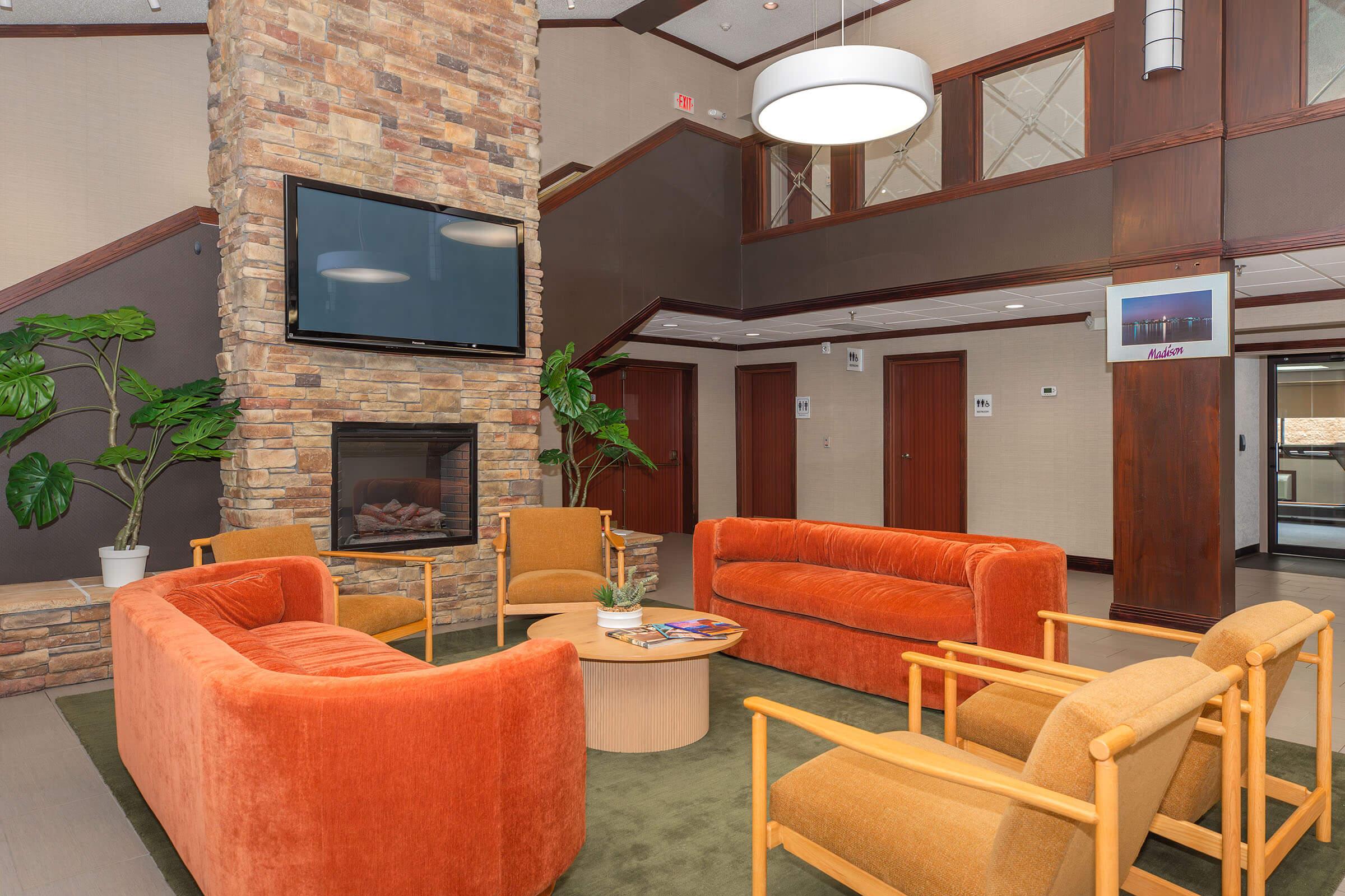 A modern lobby area featuring a stone fireplace, four orange sofas arranged around a circular coffee table, and two wooden chairs. The space is well-lit with a large window and artificial lighting, complemented by potted plants and tasteful decor, creating a welcoming atmosphere.
