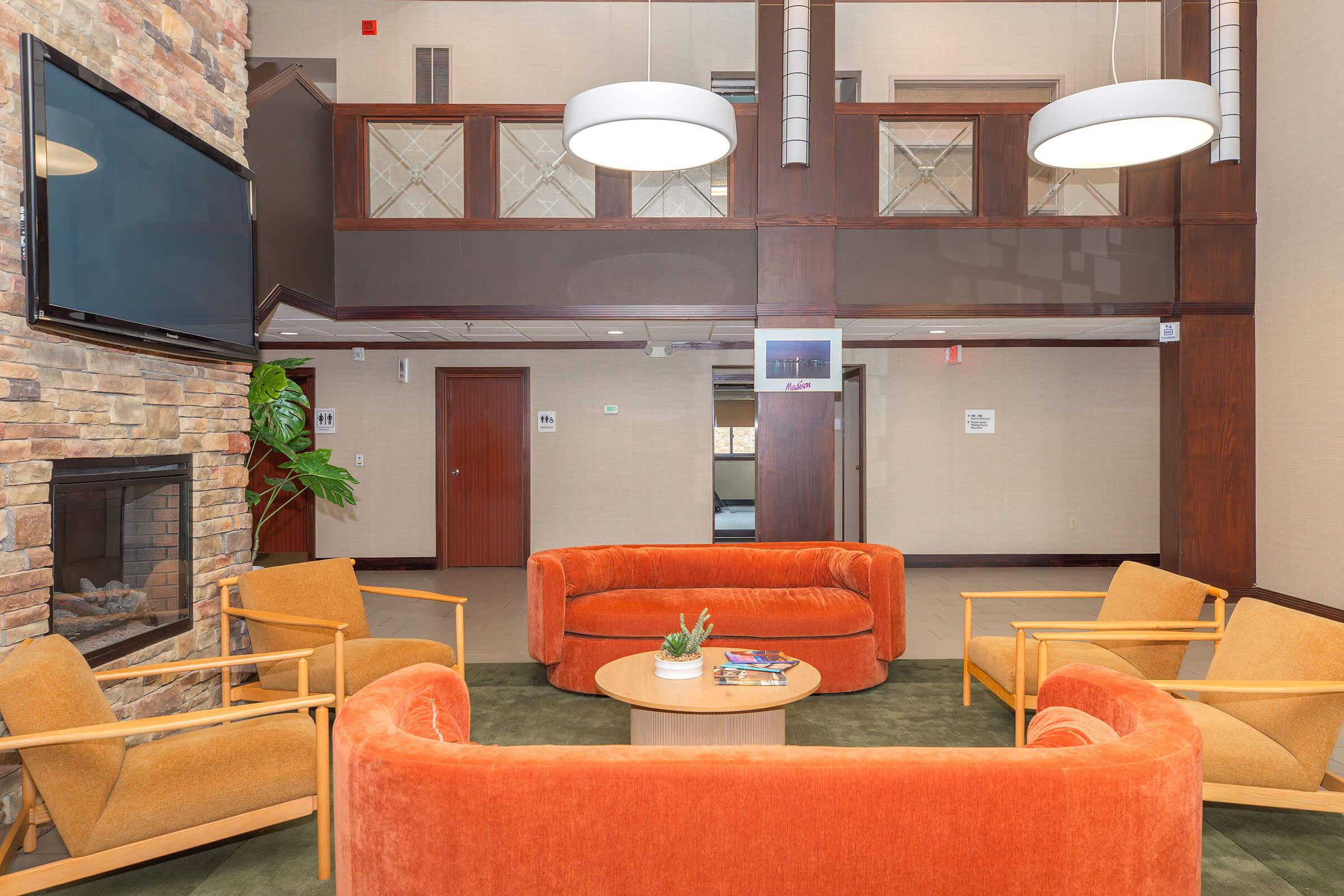 A cozy lounge area featuring orange circular sofas and wooden armchairs arranged around a round coffee table. The background includes a stone fireplace and a large TV on the wall, with a well-lit, welcoming atmosphere and modern design elements.