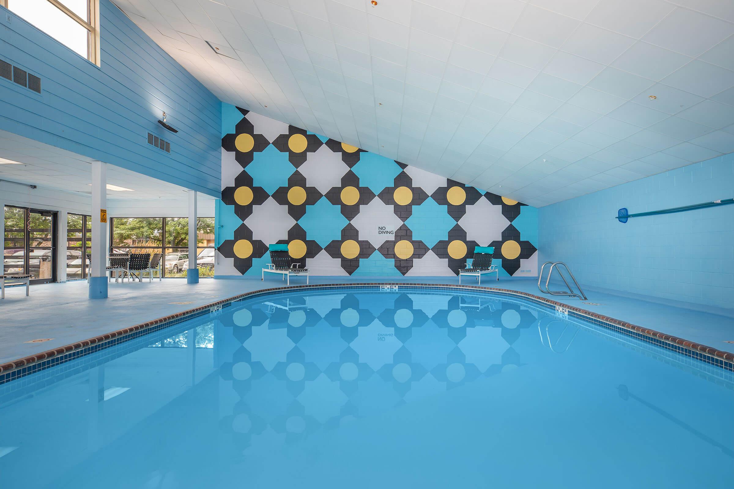 Indoor swimming pool with a geometric patterned wall featuring blue, black, and gold colors. The pool is surrounded by lounge chairs and large windows that allow natural light to fill the space. The ceiling is high and sloped, contributing to a bright and airy atmosphere.