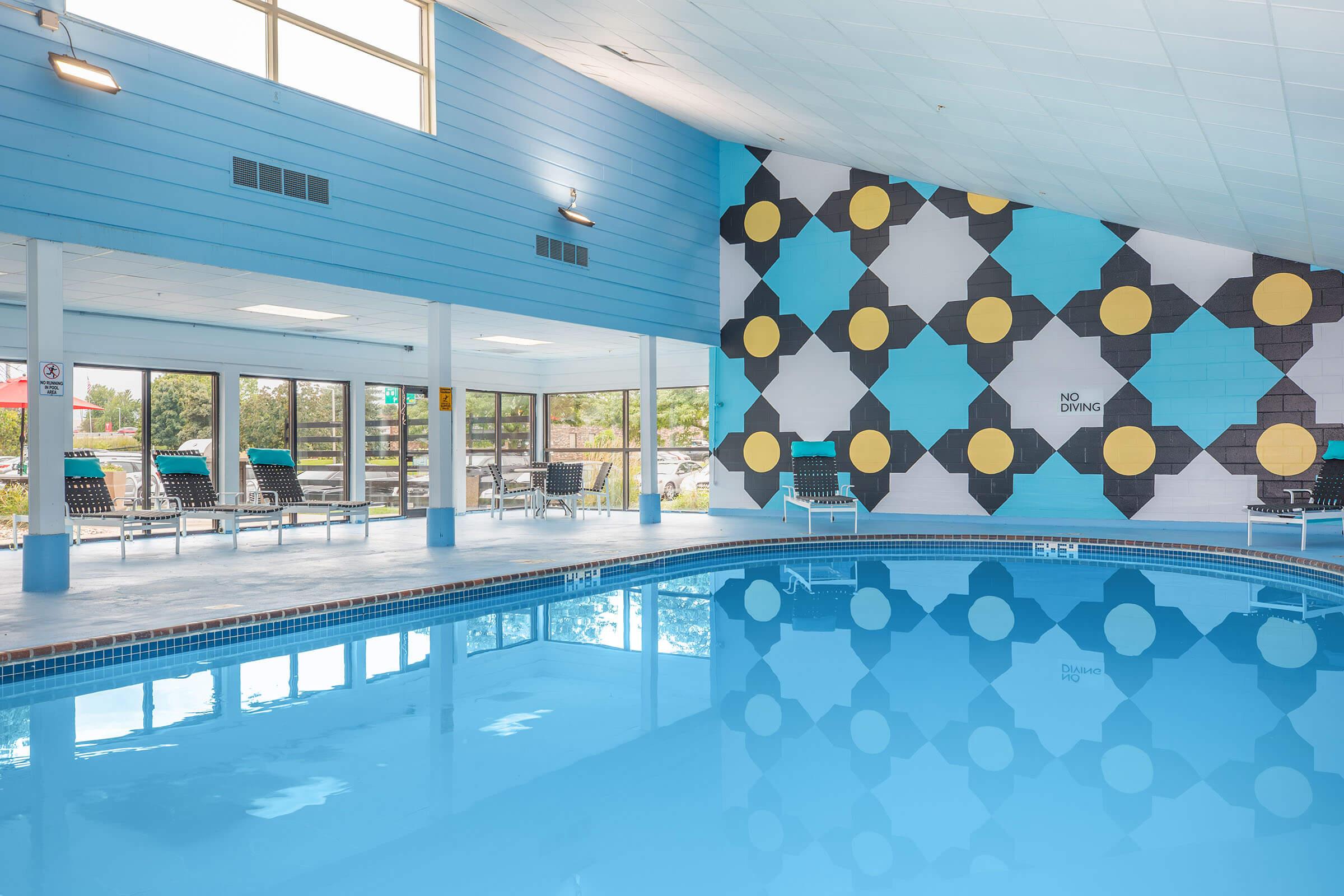 Indoor swimming pool with bright blue water and a reflective surface. The walls are painted in a combination of blue and a colorful geometric pattern featuring yellow circles. Sunlight streams through large windows, and lounge chairs are arranged near the poolside. A "No Diving" sign is visible on the wall.