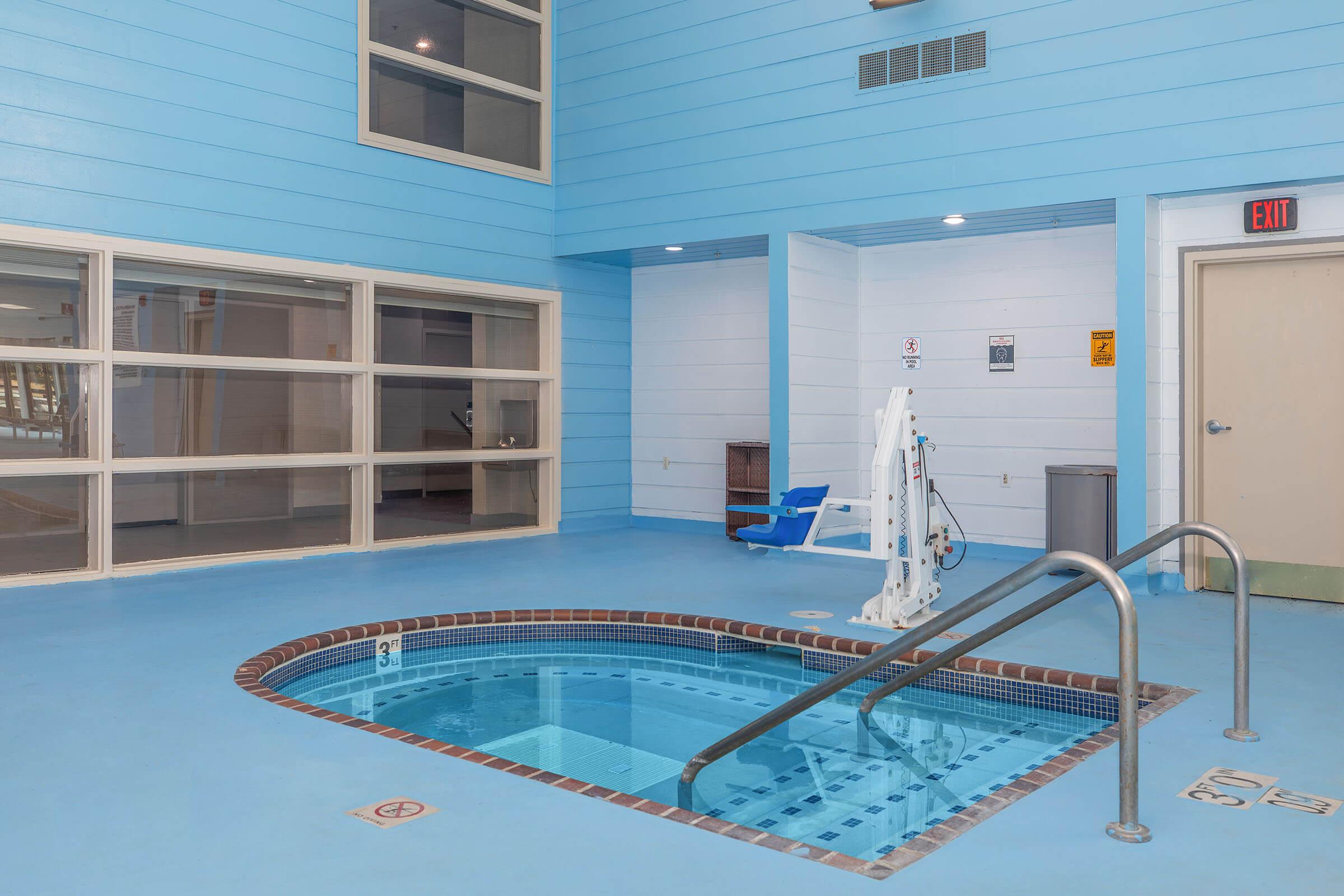 Indoor swimming pool area featuring a small, rectangular spa with a blue-tiled interior. The surrounding walls are painted light blue, and there are large windows allowing natural light. A pool lift for accessibility is visible, and safety signs are posted nearby. The floor is smooth and light-colored.