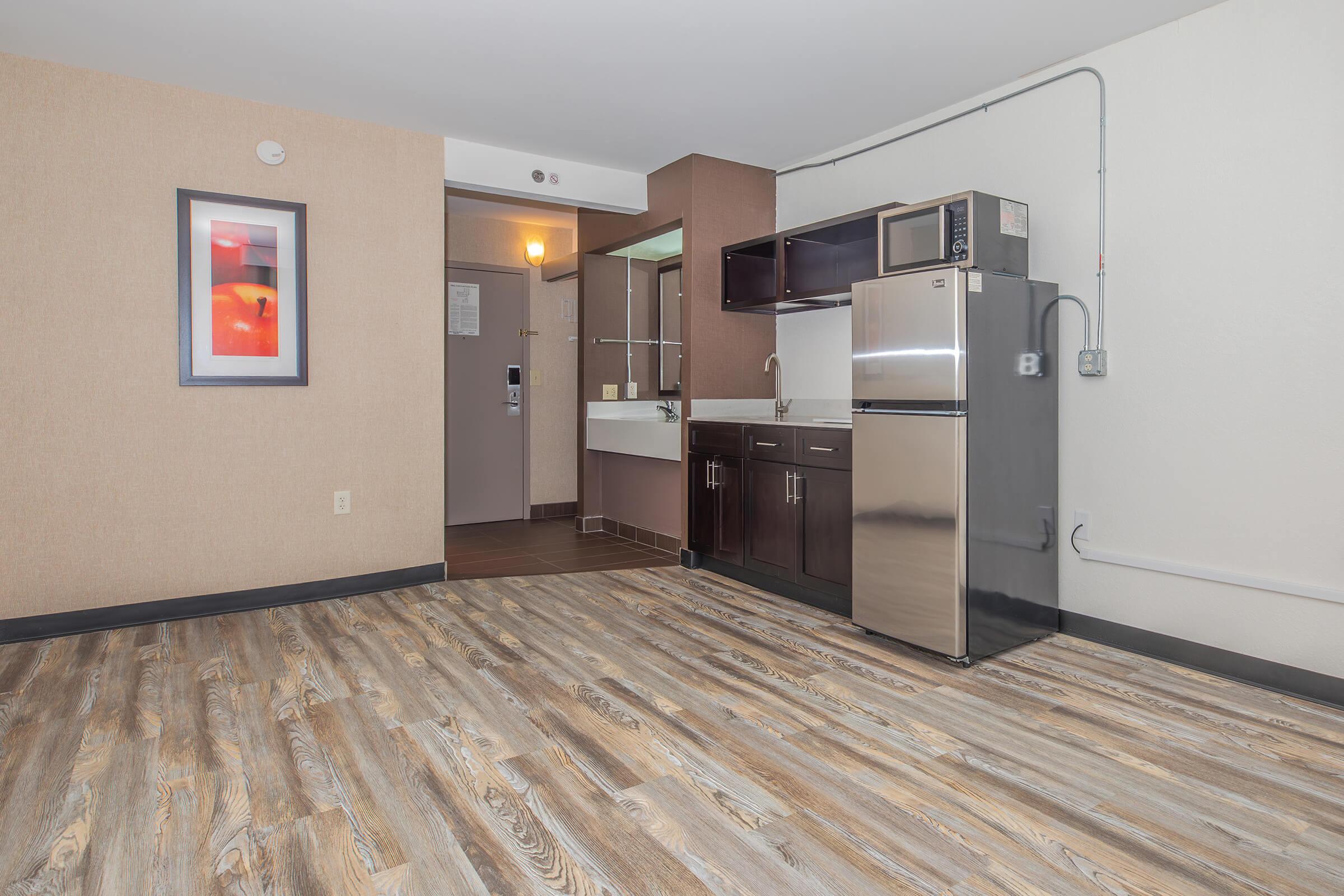 A modern studio apartment featuring a kitchenette with a stainless steel refrigerator and microwave, a bathroom area visible in the background, and wood-style flooring. The walls are a neutral color, and there is a framed piece of art on the wall.