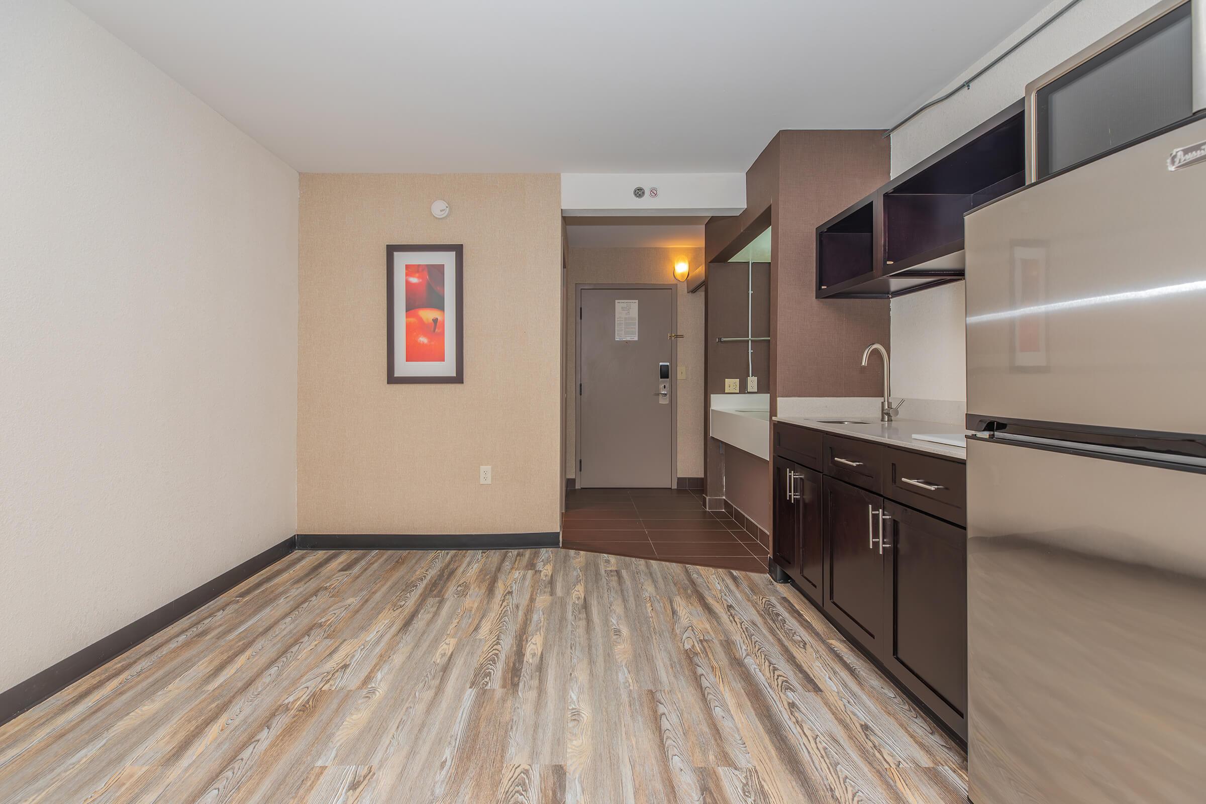 A spacious hotel room featuring a kitchenette with dark cabinetry, a stainless steel refrigerator, and a sink. The floor is modern with wood-like tiles. A bathroom area is visible with a mirrored vanity. The entrance door is seen in the background, along with a wall-mounted flat-screen TV.