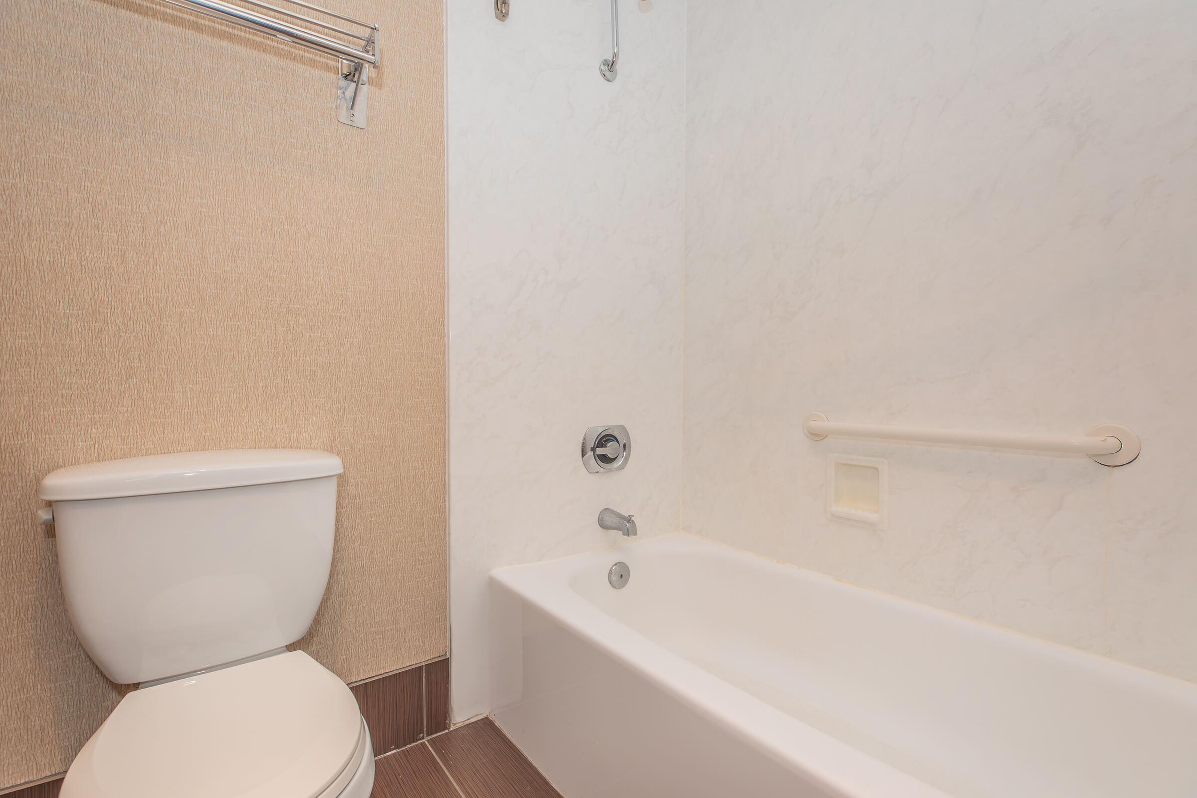 A clean bathroom featuring a white toilet and a simple white bathtub. The walls have a light, textured finish, with a grab bar next to the tub and a metal towel rack above. The overall design is minimalistic and modern.