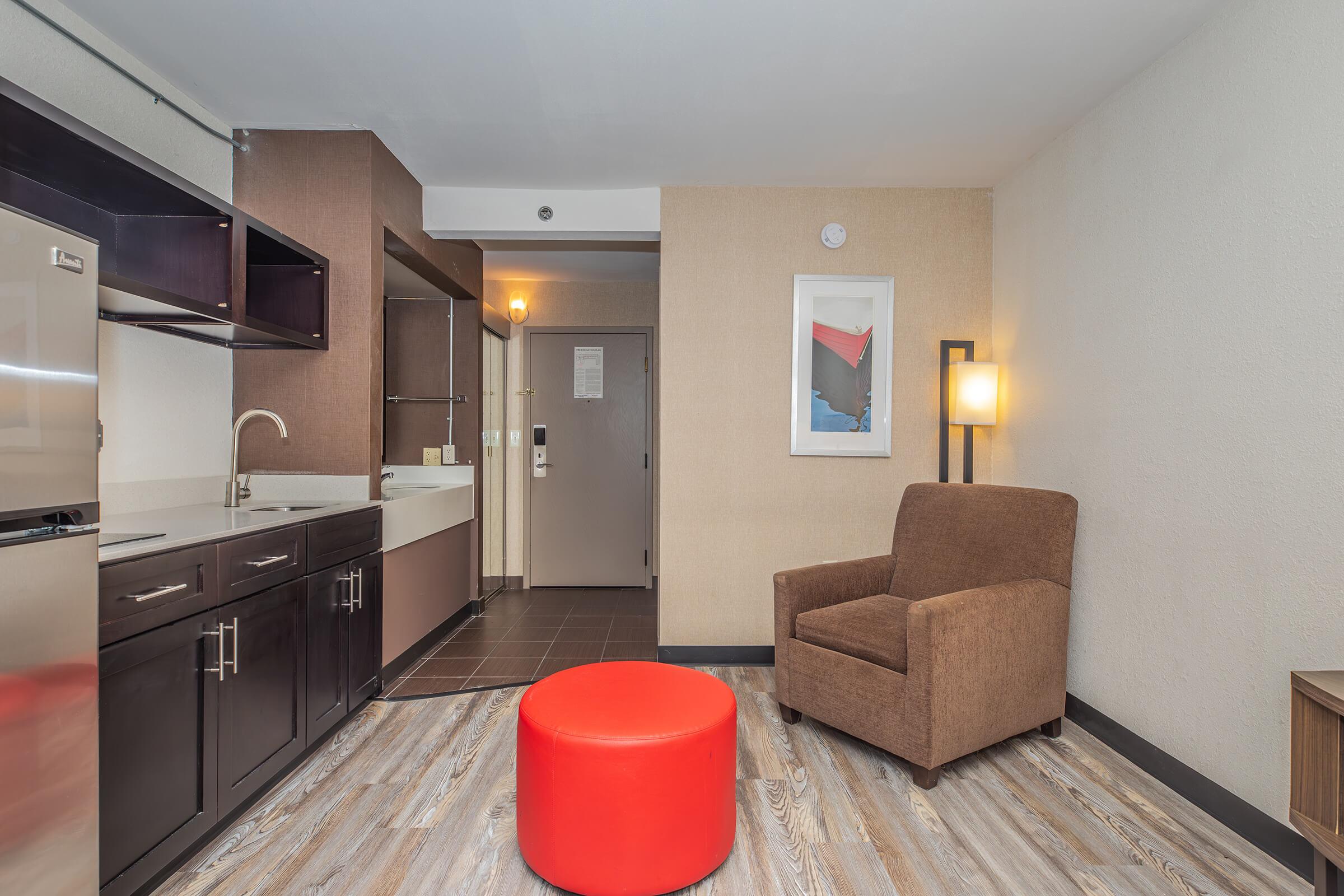 A modern hotel room featuring a kitchenette with a sink and refrigerator, a brown armchair, an orange round ottoman, a wall lamp, and artwork. The entrance door is visible in the background, along with a bathroom area. The flooring is a light wood design, creating a cozy atmosphere.