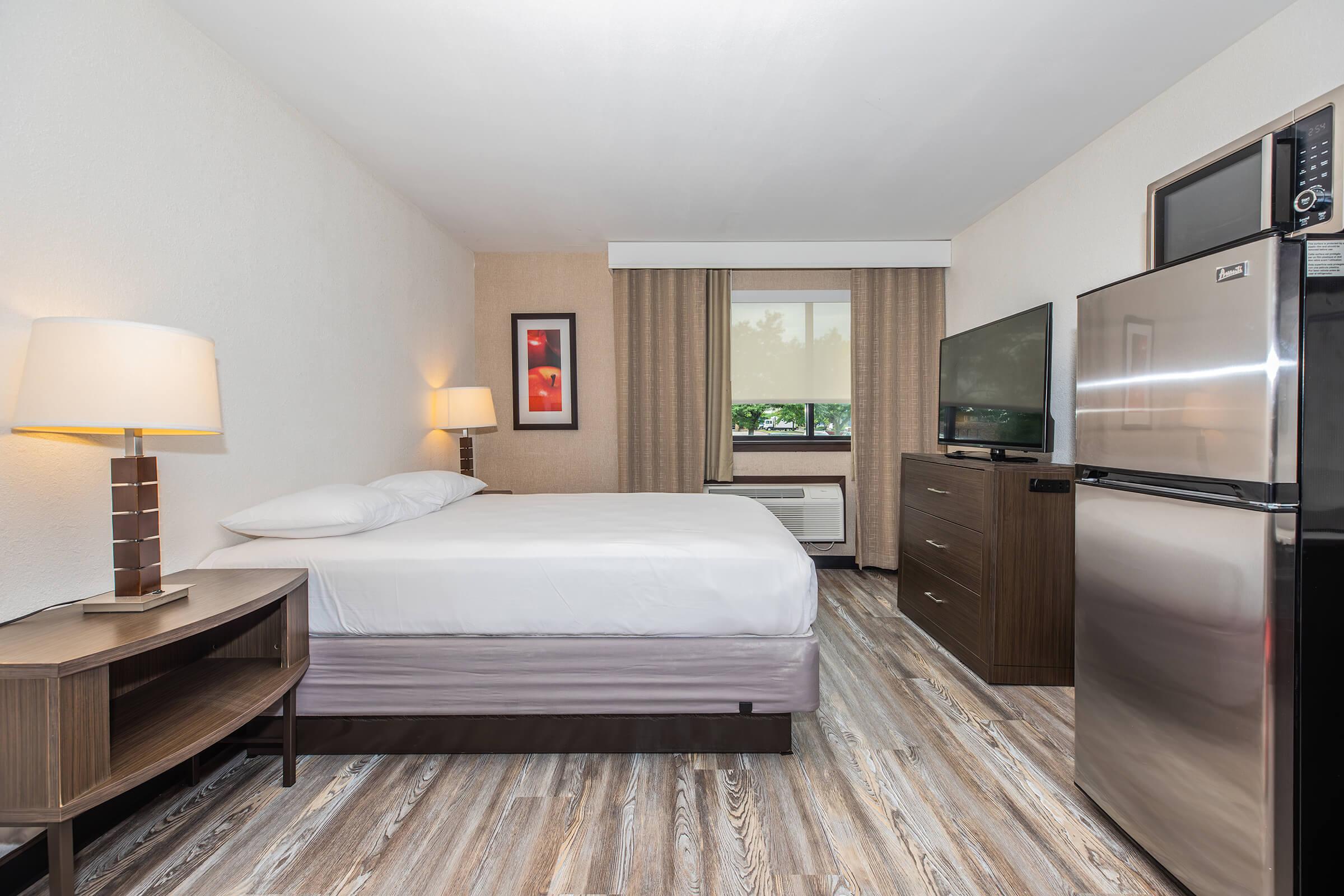A bright, modern hotel room featuring a queen bed with white linens, a bedside lamp, a flat-screen TV on a dresser, an air conditioning unit, and a mini-fridge. Natural light enters through a window covered with light curtains, and the flooring is a wood-like laminate.