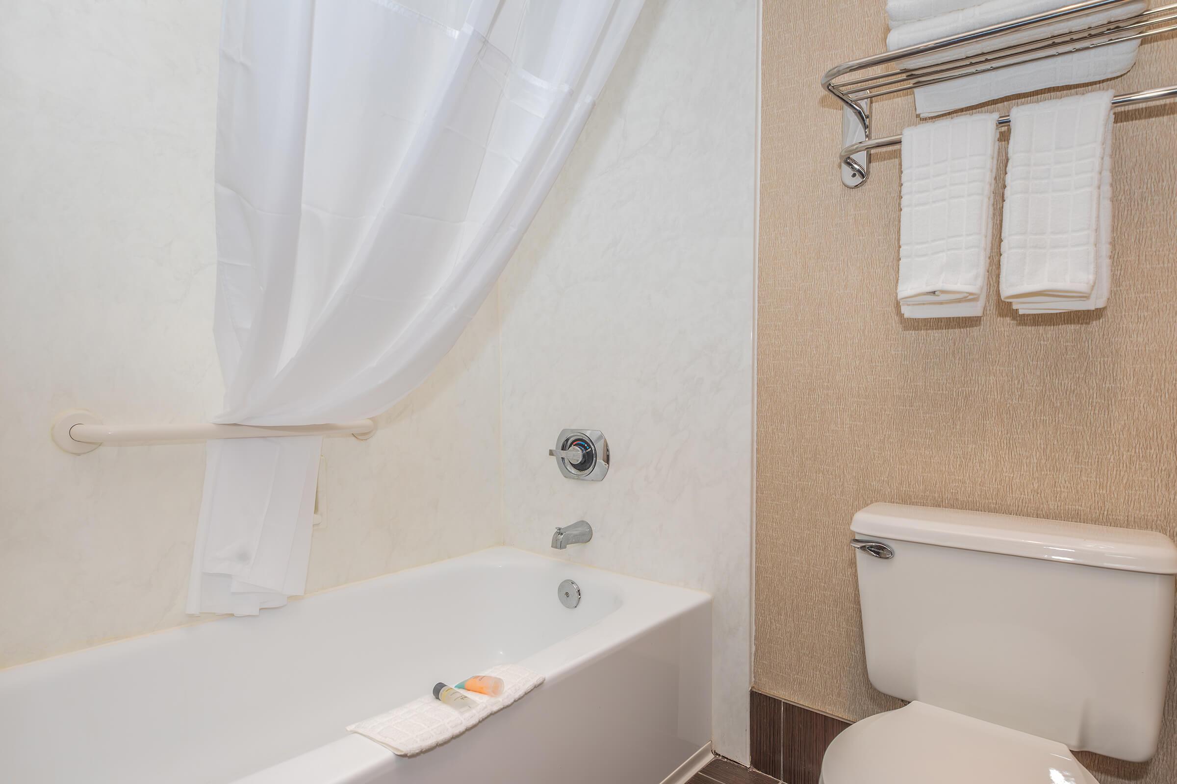 A bathroom featuring a bathtub with a white curtain, a towel rack with folded towels, and a toilet. The walls are light-colored, creating a clean and modern appearance.
