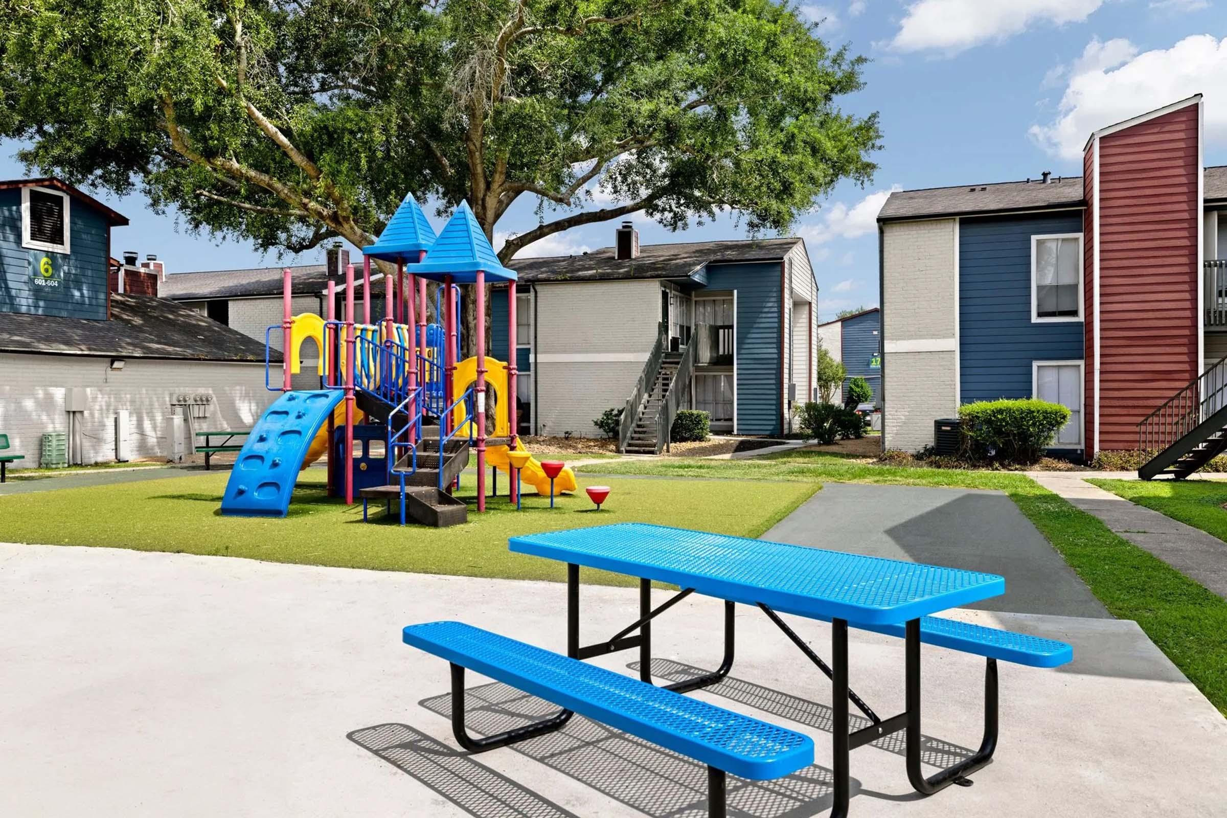 Colorful playground equipment with slides and climbing structures surrounded by green grass, a picnic table in blue, and apartment buildings in the background under a sunny sky with a few clouds.