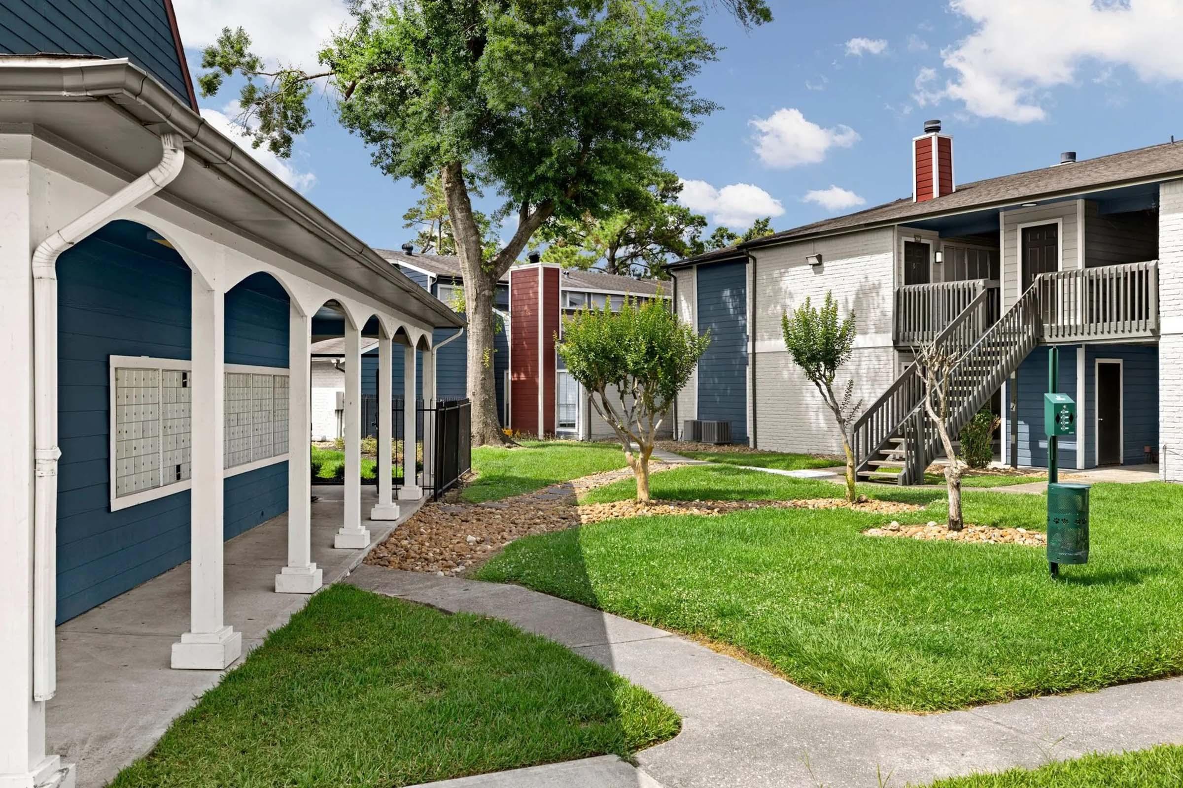 A landscaped area of an apartment complex featuring a pathway, greenery, and buildings with balconies. One building is blue with white trim, while another has gray siding. There are trees and small shrubs amid the grassy space, creating a pleasant communal outdoor environment.