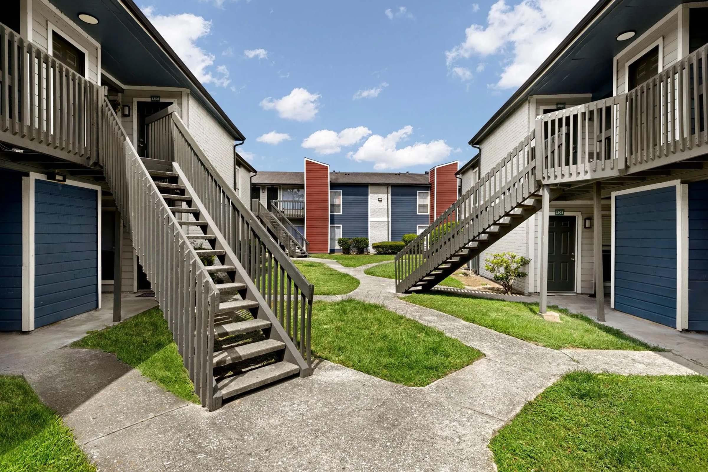 A view of an apartment complex featuring two-story buildings with blue and red accents. There are staircases leading to the upper levels on either side, and a grassy courtyard with a path running through it. The sky is partly cloudy, creating a bright and welcoming atmosphere.