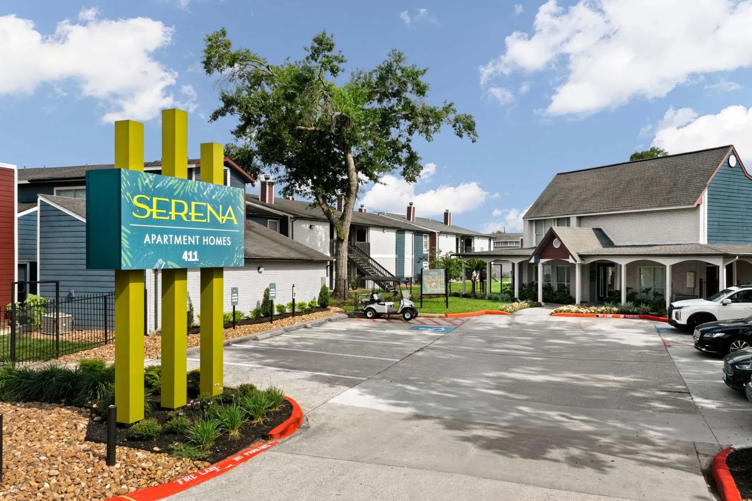 Sign for Serena Apartment Homes with a modern design, surrounded by landscaped greenery. In the foreground, a paved parking area features a golf cart and two parked vehicles. The buildings have a mix of blue and gray colors, set against a bright blue sky with scattered clouds.